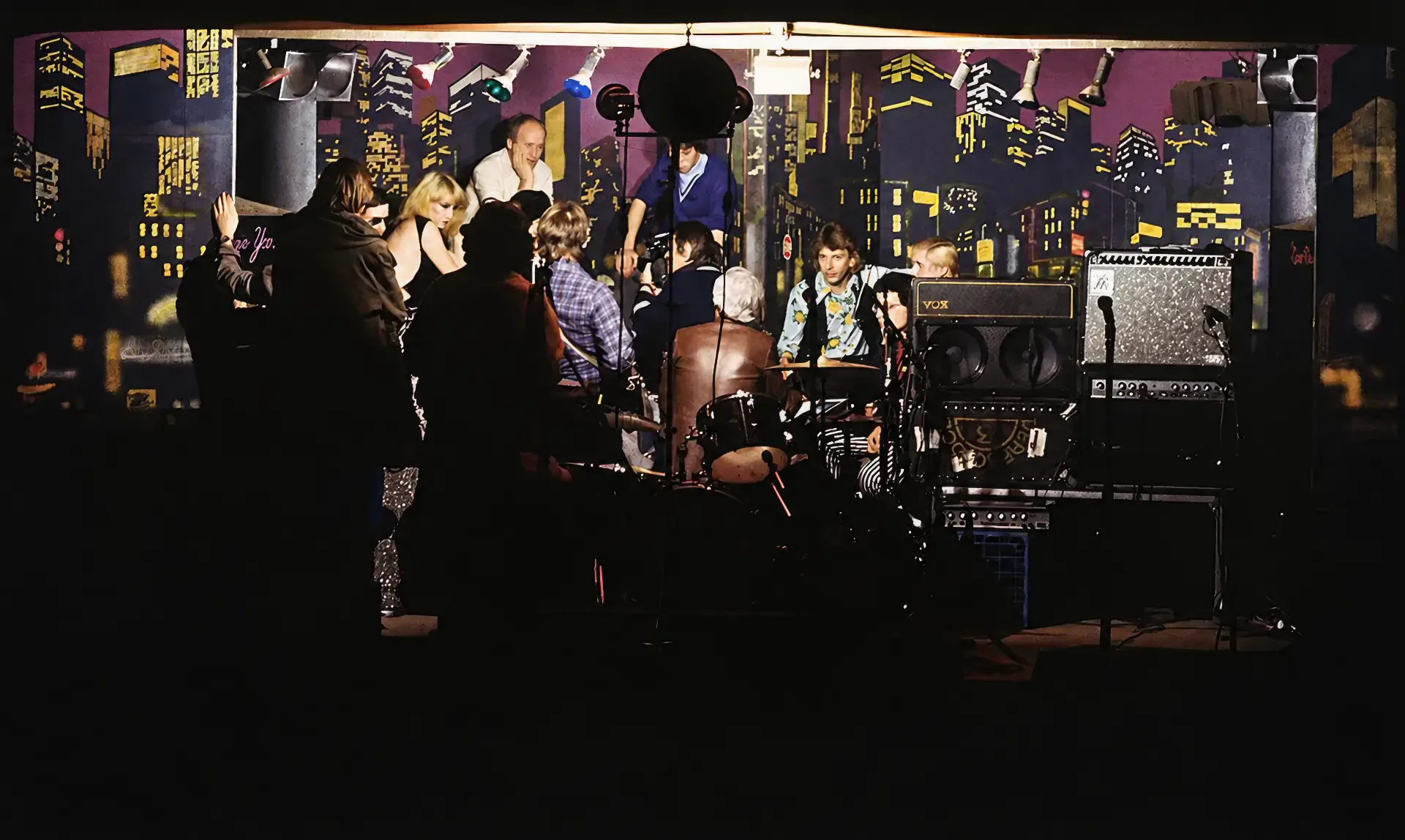 Photographie couleur, format horizontal. En 1977, avant leur concert au Canotier, les musiciens de Deaf School répondent à la presse au milieu des amplificateurs Vox, des fûts et des projecteurs. Tout autour, le décor peint par Aldo Gigli : une ville nocturne aux façades violettes, fenêtres jaunes et enseignes au néon, qui enveloppe la scène comme un décor de cinéma.