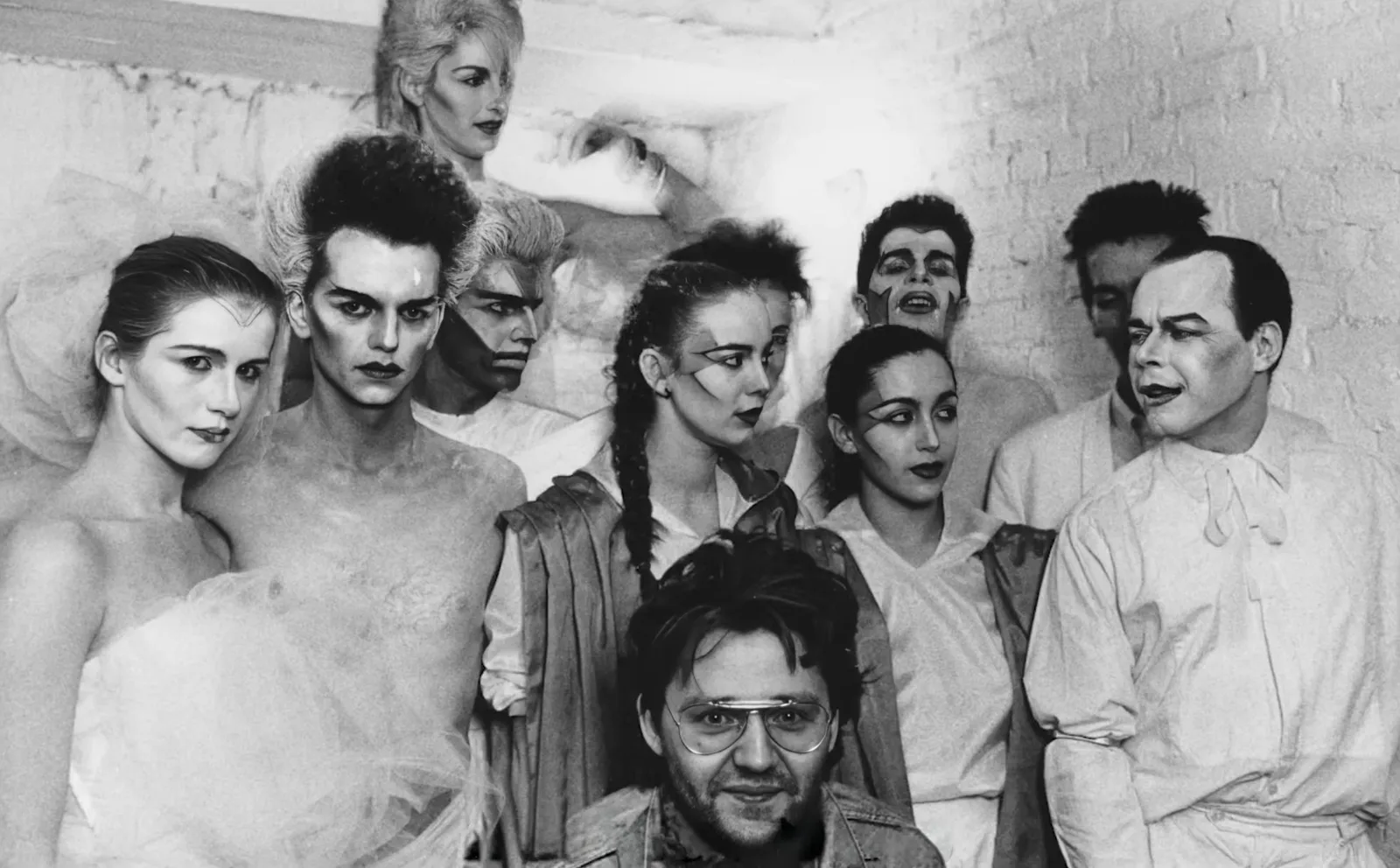 Photographie noir et blanc, format horizontal. Les danseurs et performeurs du Mirano Continental réunis contre un mur de brique : visages maquillés en blanc, traits graphiques noirs, costumes de théâtre. À droite, Bruno Bulté, qui fut aussi président de la Ligue d'Improvisation Belge, dont les matchs avaient lieu au Mirano. Années 1980.