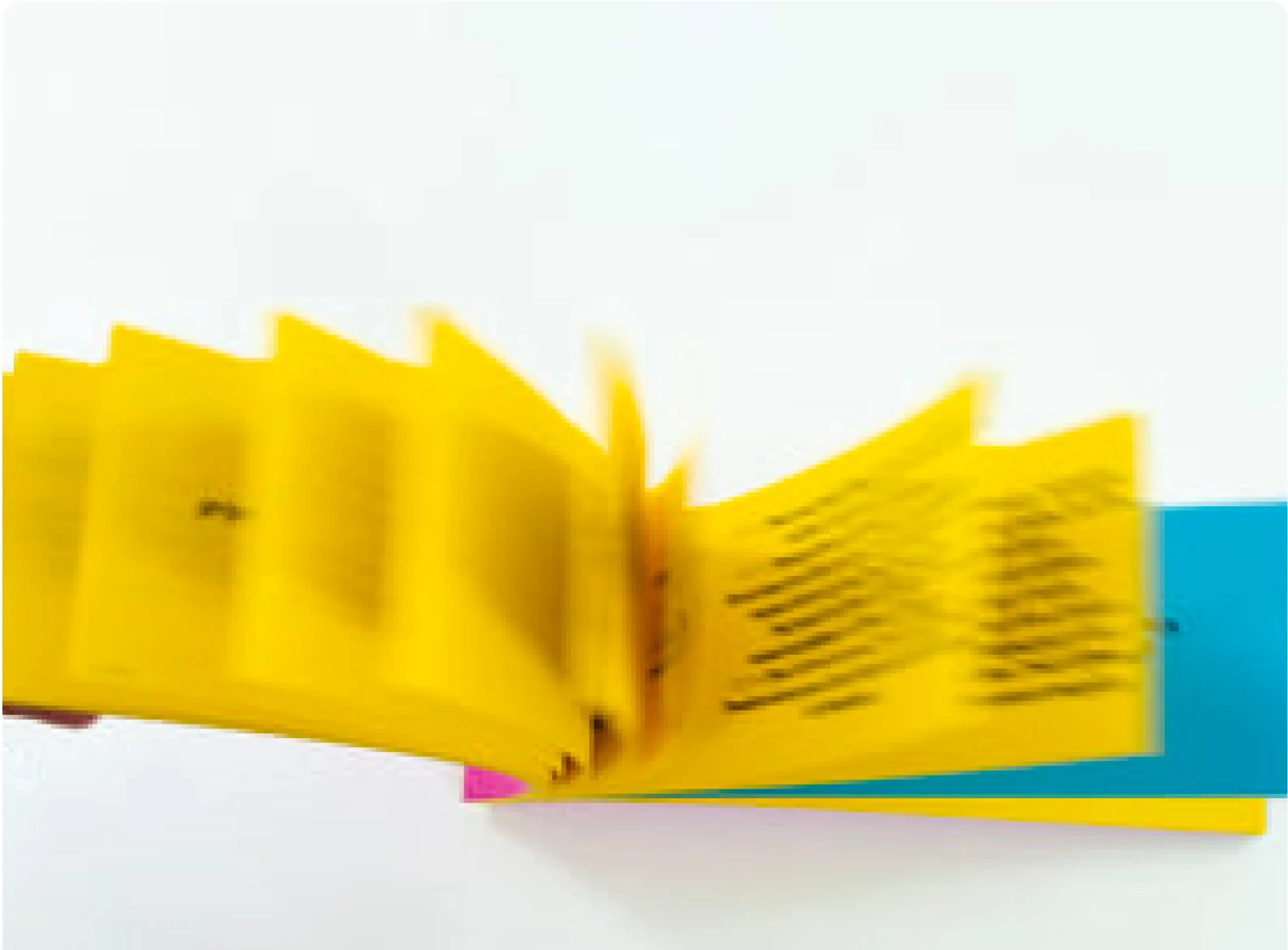 Photographie de l'éditeur : le carnet ouvert en plein mouvement, pages jaunes floutées par le geste, tranches turquoise et magenta posées dessous. Image de la fiche produit d'afterclub.be, où le carnet est vendu 10 euros.