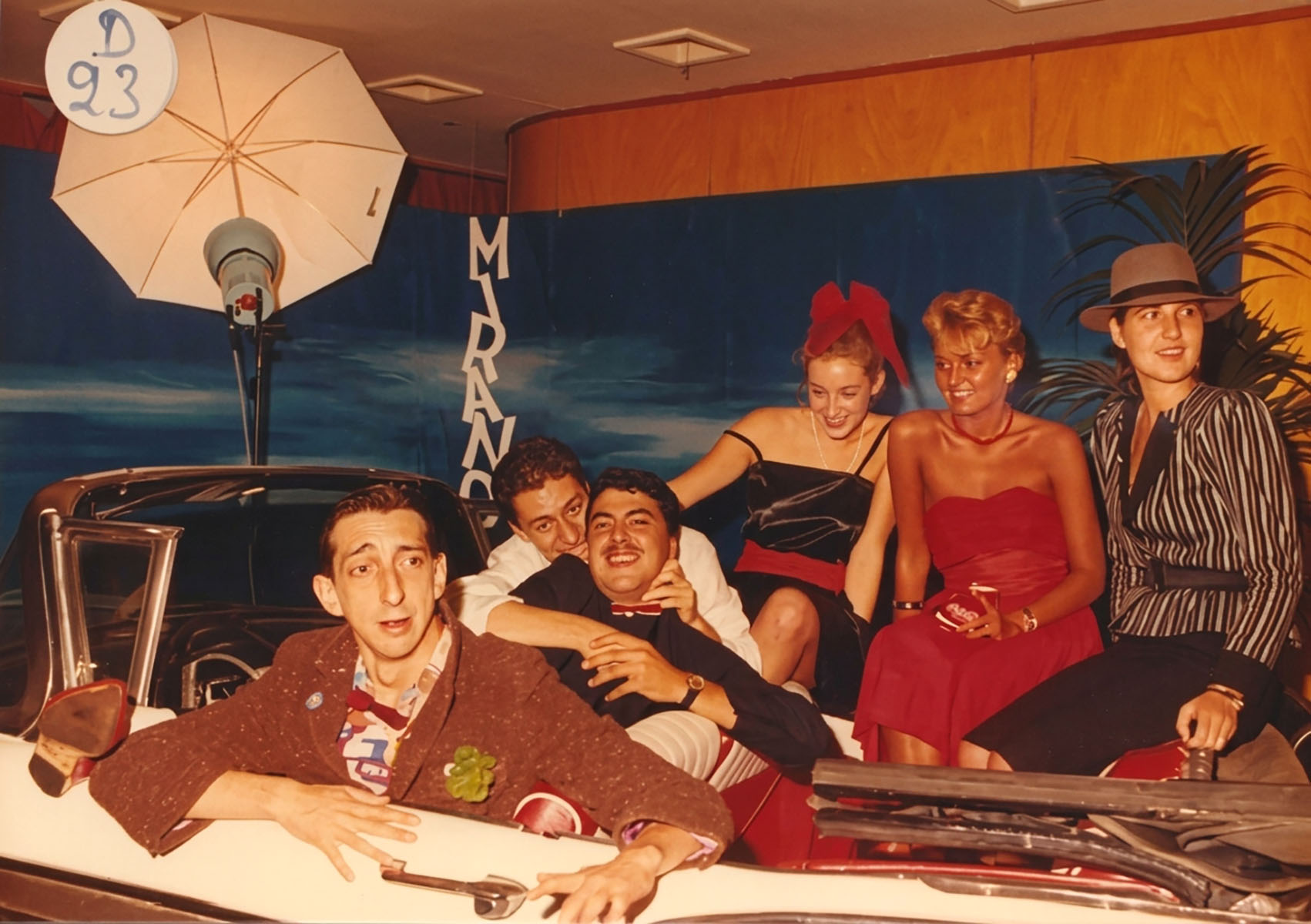 Photographie couleur, format horizontal. Même séance de 1989 dans le hall du Mirano Continental : Dan Mac Roll, Santino Del Paradiso Trovato et Riton Liebman, le Trio Infernal, et leurs invitées rient à bord de la Cadillac, entre parapluie réflecteur de studio et décor marin peint à la main.