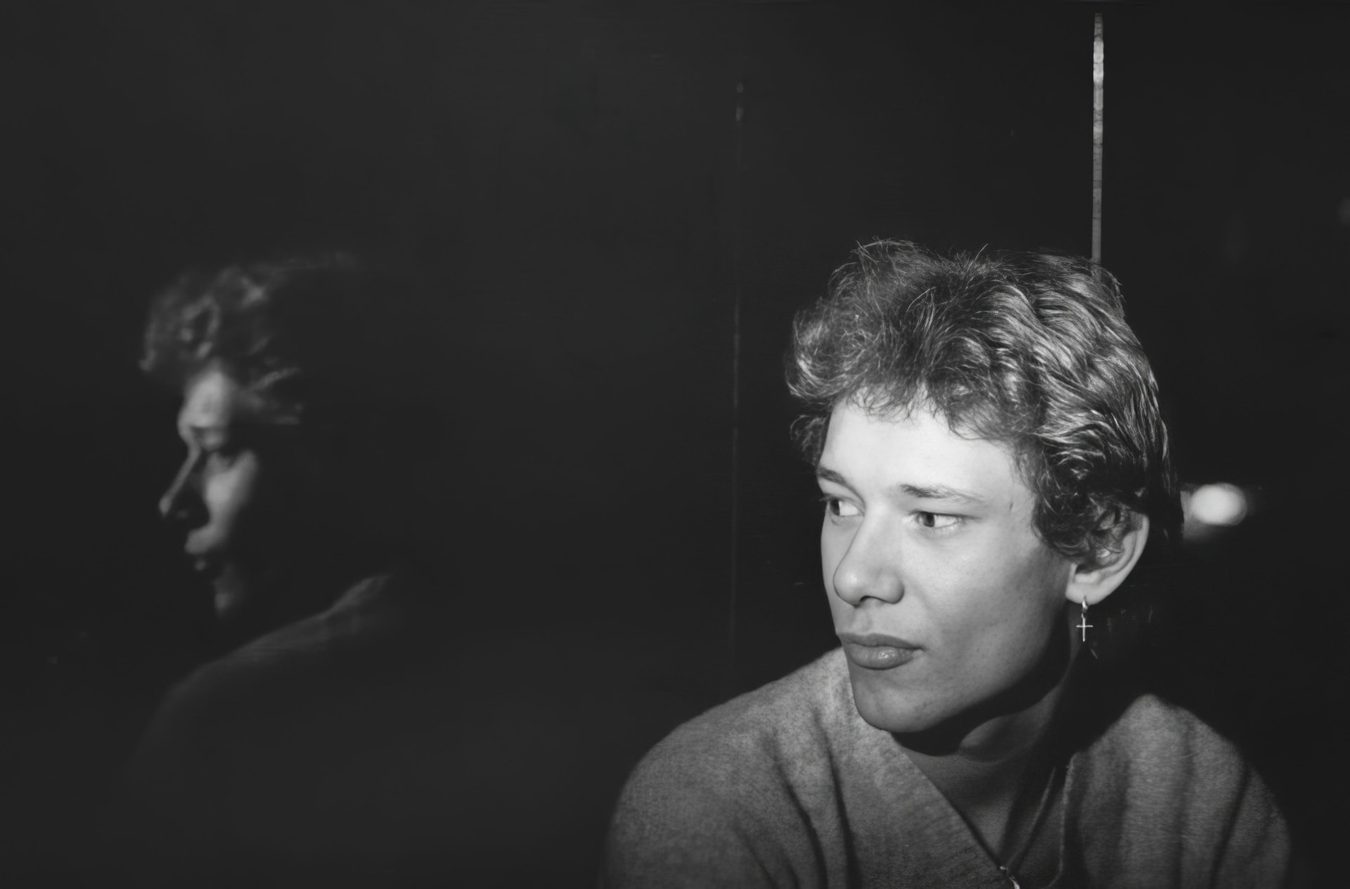Photographie noir et blanc, format horizontal. Nanard, figure des nuits du Canotier, photographiée au club en 1977, une croix à l'oreille, dans la pénombre de la salle.