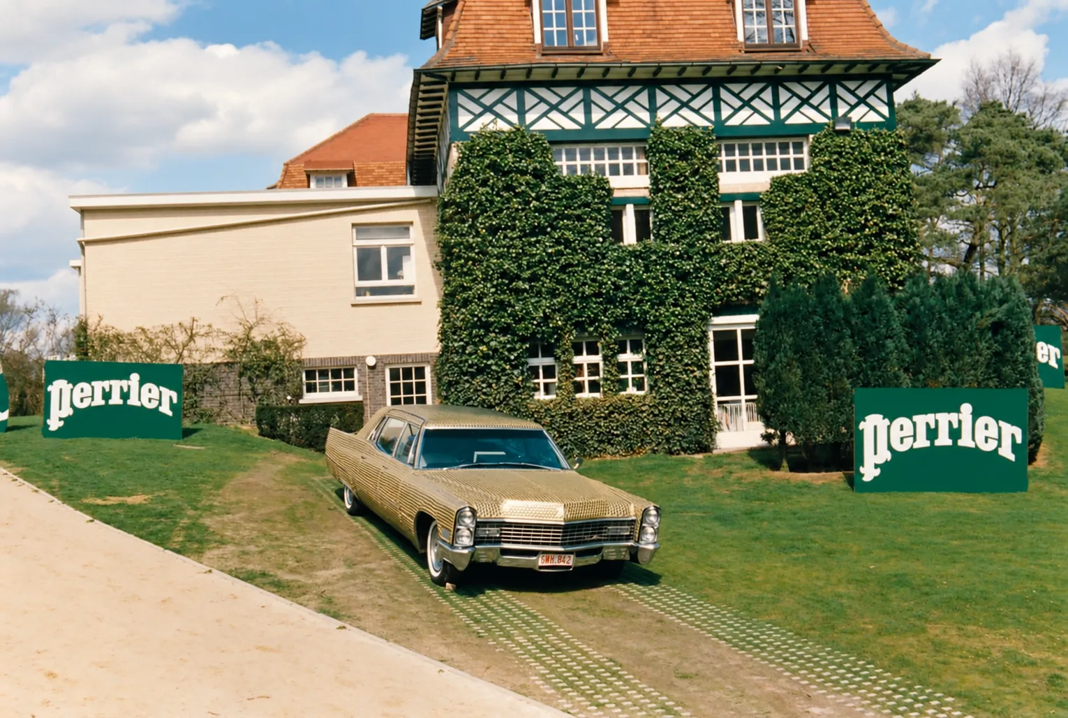 Photographie couleur, format horizontal. Sur une pelouse, devant une villa à colombages couverte de lierre taillé, une berline américaine des années 1960 entièrement habillée de capsules dorées, plaque belge GMH 842. De part et d'autre, de grands panneaux verts frappés du logo Perrier en lettres blanches. La voiture, recouverte capsule par capsule, transforme une carrosserie ordinaire en objet publicitaire de collection.