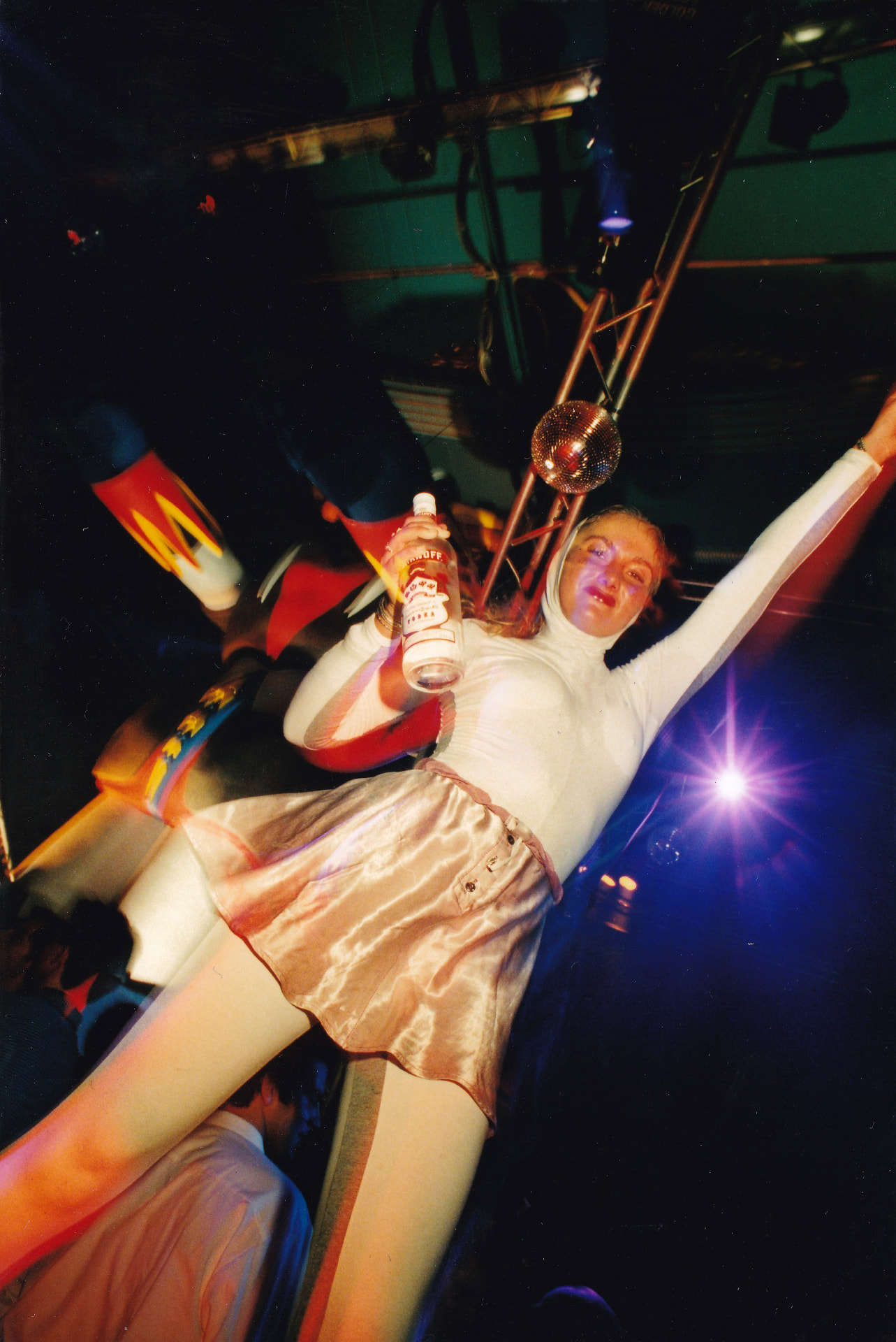 A dancer in a white bodysuit on the podium, a bottle of Smirnoff vodka (product placement before its time) in hand, beneath the mirror ball.