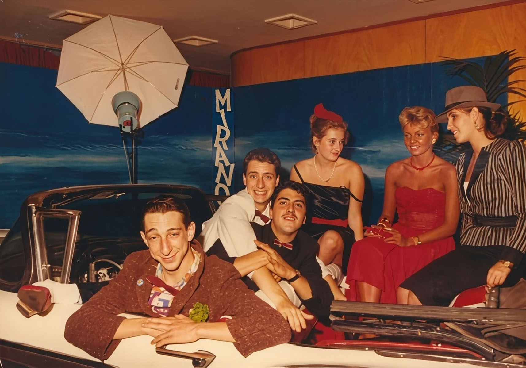 Photographie couleur, format horizontal. En 1989, Dan Mac Roll, Santino Del Paradiso Trovato et Riton Liebman, le Trio Infernal, posent avec leurs invitées à bord de la Cadillac installée dans le hall d'entrée du Mirano Continental, sous le parapluie réflecteur du studio photo et devant le décor bleu nuit au logo vertical du club.