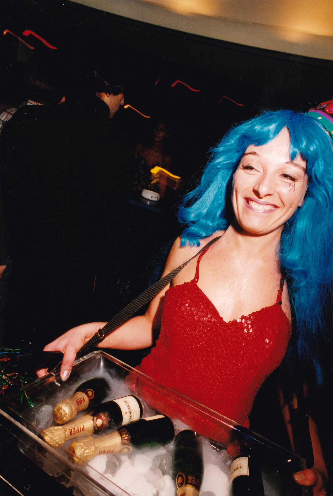 Piper-Heidsieck hostess in a blue wig and red sequin dress, carrying a tray of bottles on ice.