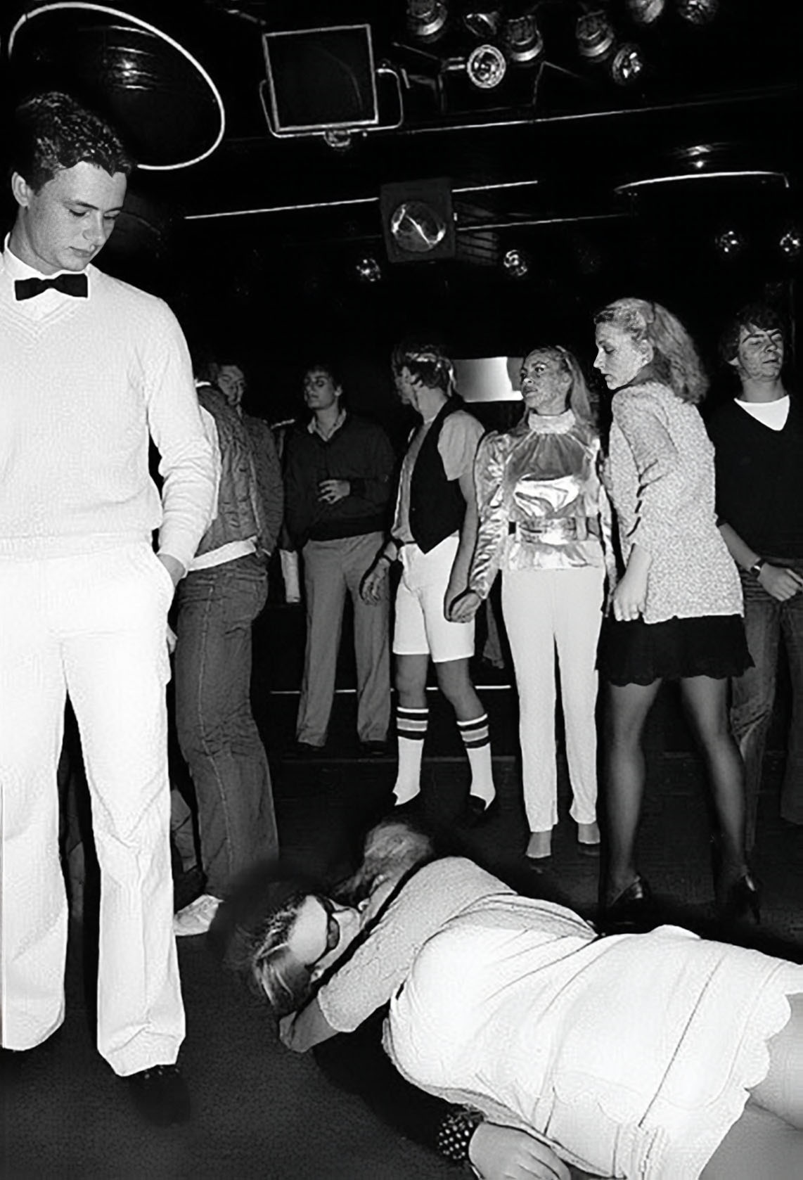 Photographie noir et blanc, format horizontal. Vue d'ensemble de la salle du Mirano Continental : un homme en costume blanc allongé au premier plan, d'autres clients debout ou assis, le bar et les lumières de la discothèque en arrière-plan.