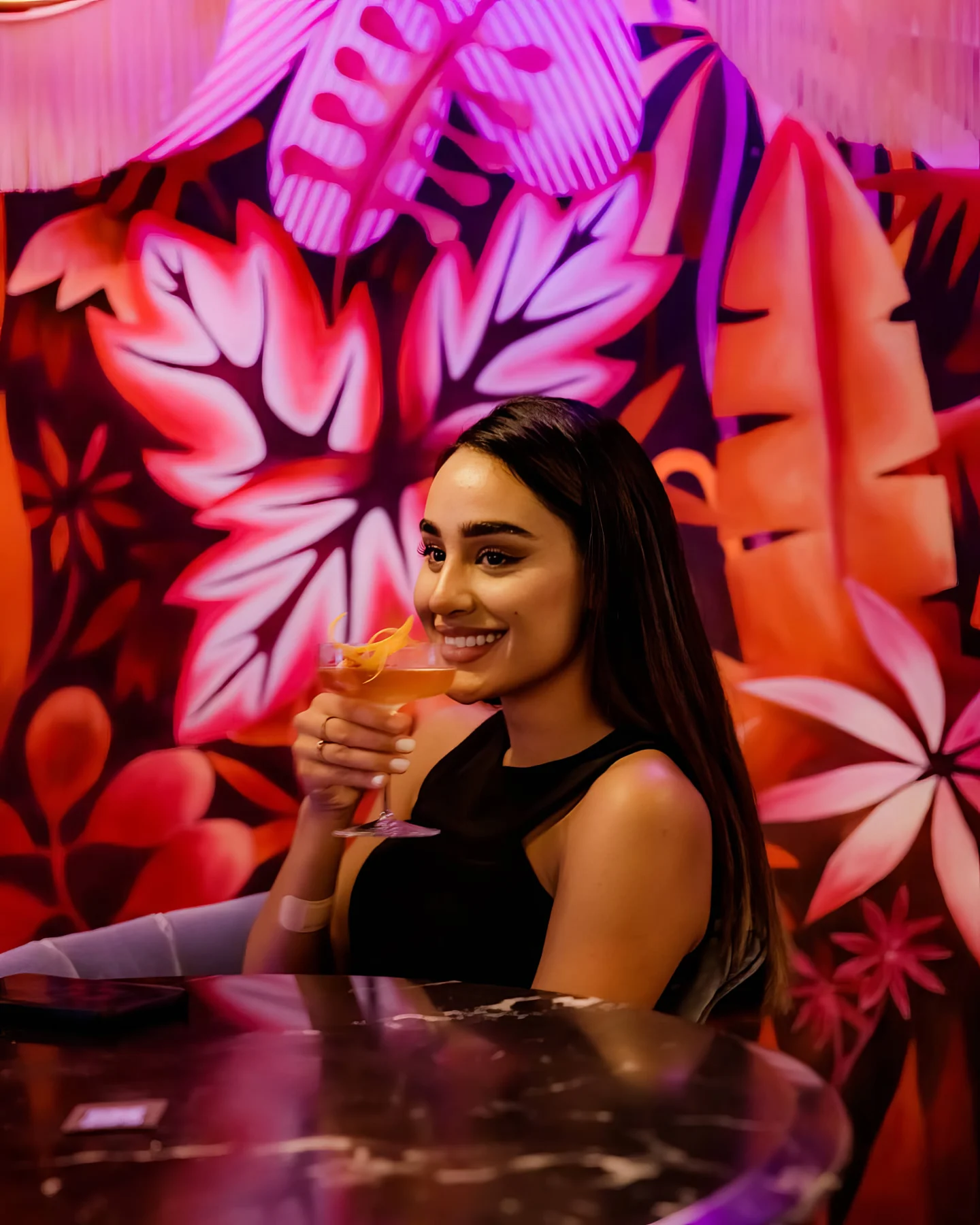 Une cliente pose son verre devant la partie la plus rose de la fresque : grandes palmes ouvertes, fleurs stylisées, rouges chauffés par la lumière. Le décor fonctionne comme un fond de portrait, ce qu'Aldo Gigli a toujours cherché.