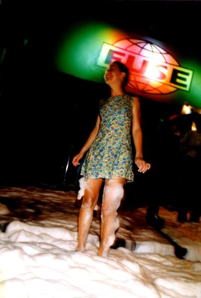 A dancer in a flowered dress, barefoot in the foam, beneath the illuminated Fuse logo.