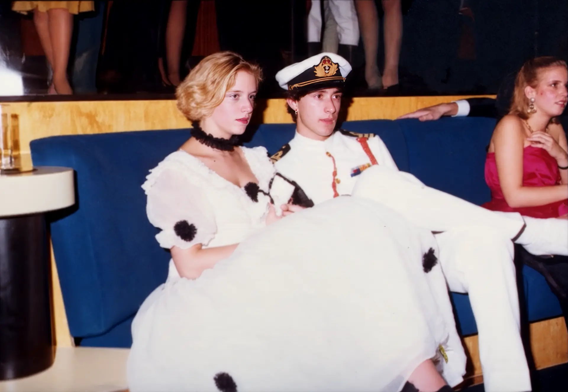Photographie couleur, format horizontal. Au Mirano Continental, un couple en costume de scène : elle en robe blanche à pompons noirs et collier de plumes, lui en uniforme de marin blanc avec casquette d'officier. Assis sur le canapé bleu emblématique de la salle, ils incarnent l'esprit théâtral et décalé des soirées à thème du club dans les années 1980.