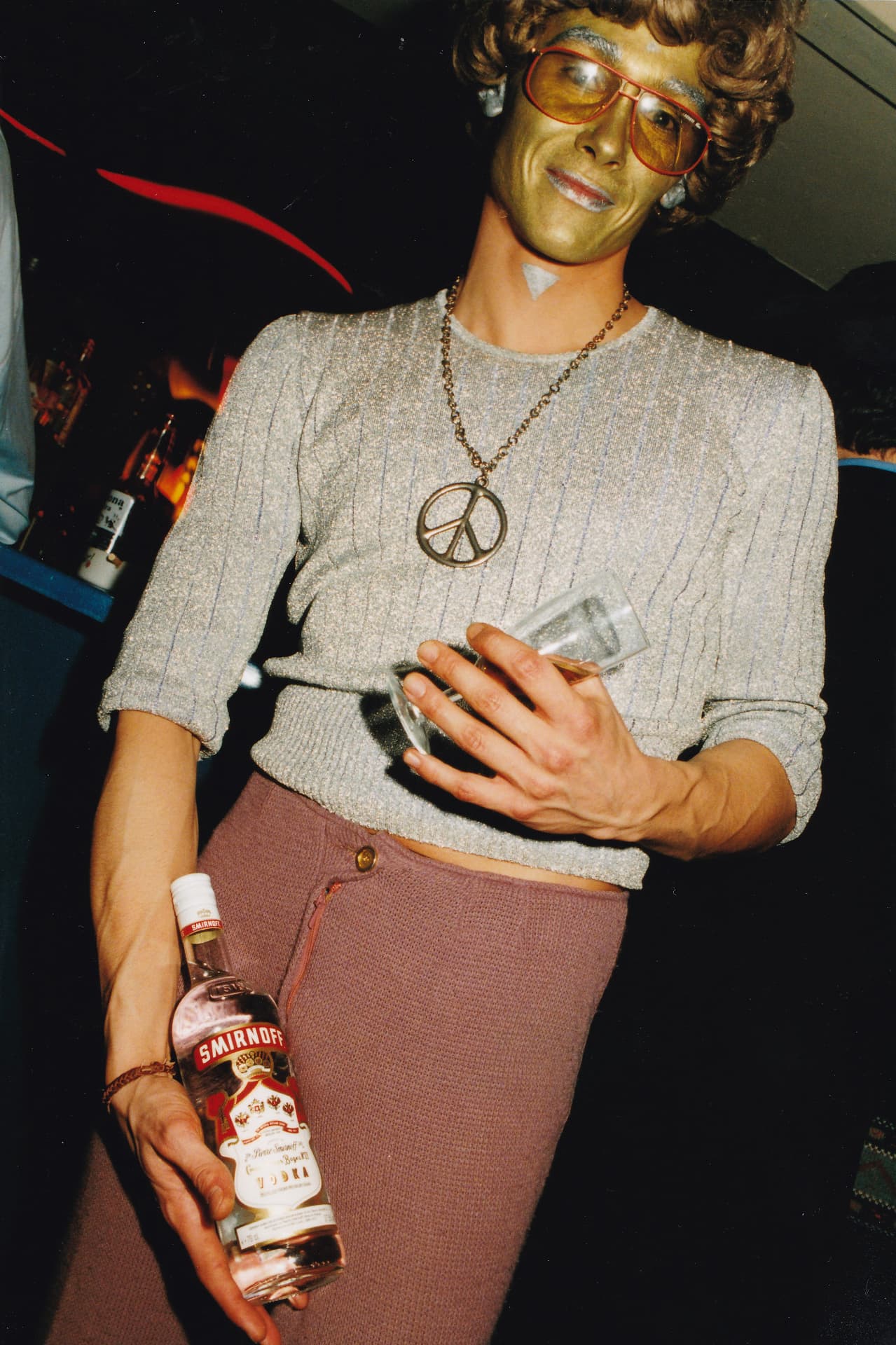 Portrait at the bar: gold-painted face, tinted glasses and a peace-and-love pendant.