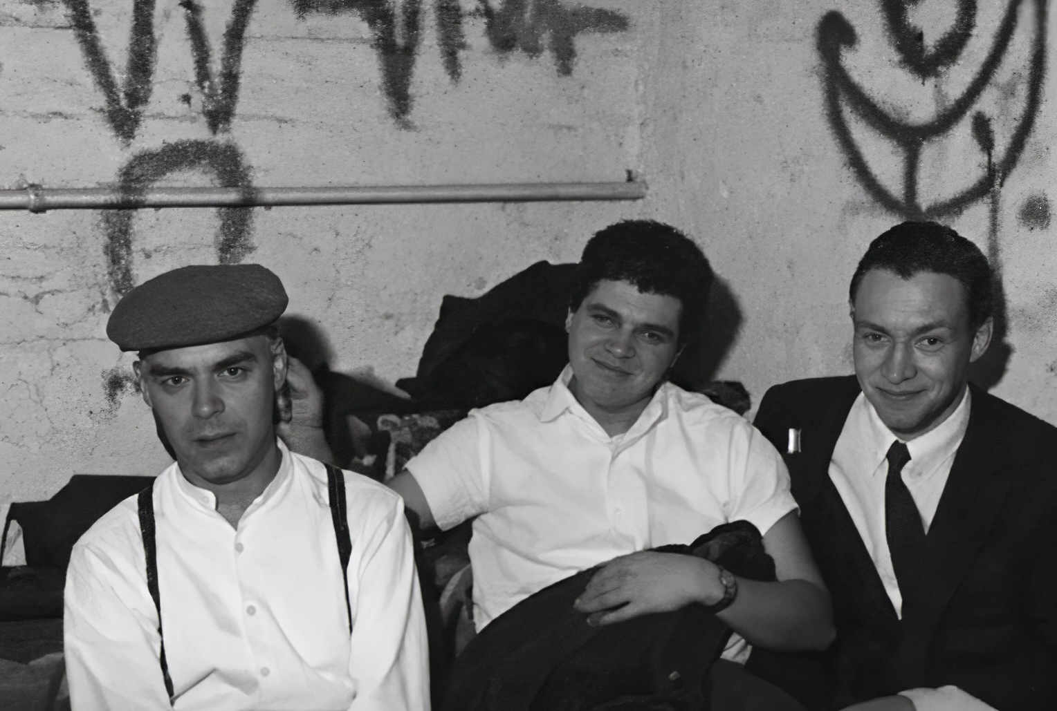 Photographie noir et blanc, format horizontal. Trois hommes posent devant un mur tagué du Mirano Continental ; leurs styles mêlent béret, bretelles et costume, entre street culture et tenue de soirée.
