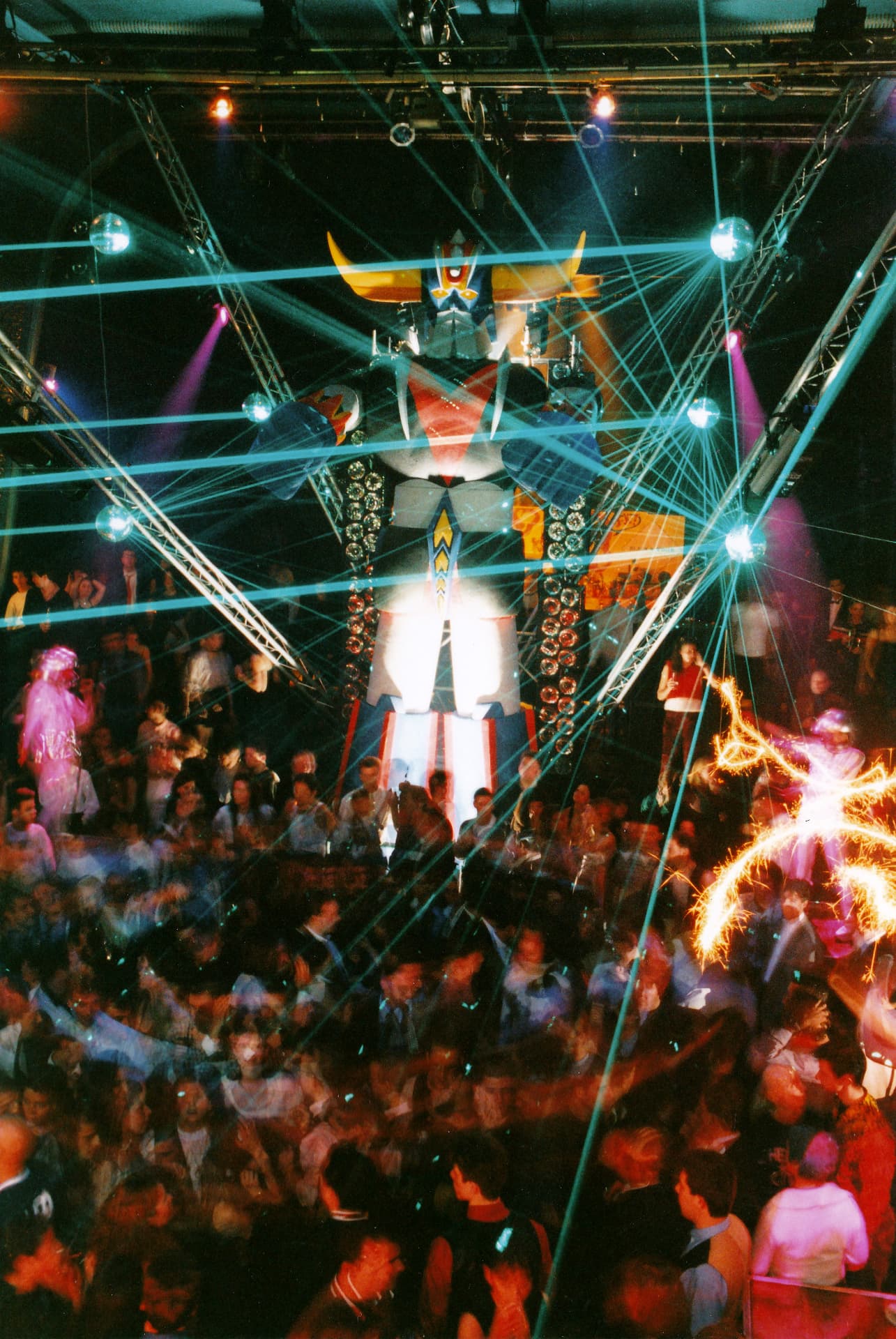 High-angle view of the room: the DJ launches the « Rétrolaser » sample and, to the second, the lighting crew fires the beams above the crowd, mirror balls and the Goldorak at the centre of the set-up.