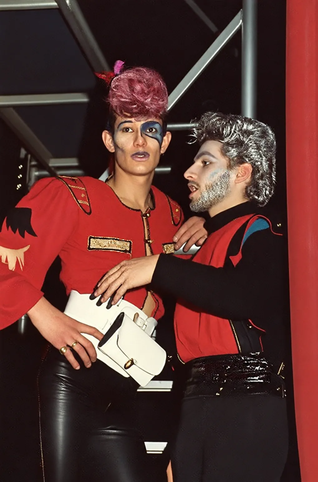 Photographie couleur, format vertical. Dans les structures métalliques du club, deux silhouettes new wave en veste rouge à épaulettes dorées et pantalon de cuir noir : coiffures sculptées rose et argent, visages maquillés au trait bleu. Nanard et Marco Rolland, pilier du Mirano et futur directeur artistique de l'agence KCC.