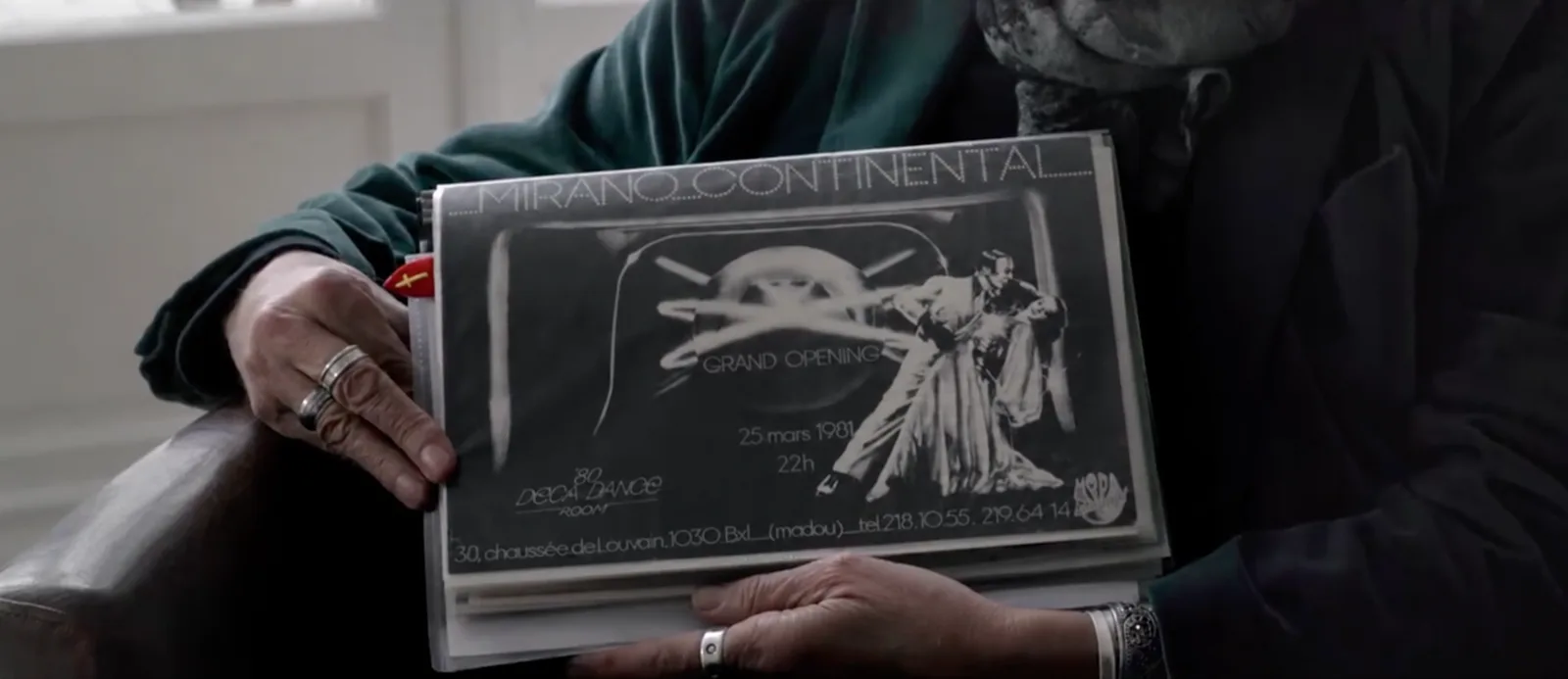 Image extraite du reportage : Aldo Gigli tient, sous pochette plastique, le visuel original de la carte de membre du Mirano Continental, pièce rare conservée depuis 1981. On y lit le lettrage en points de lumière « Mirano Continental », la mention « Grand Opening », la date « 25 mars 1981, 22h », le logo « '80 Deca Dance Room », l'adresse « 30, chaussée de Louvain, 1030 Bxl (Madou) » et les numéros de téléphone, avec le couple de danseurs en noir et blanc à droite et le logo Moda Concept.