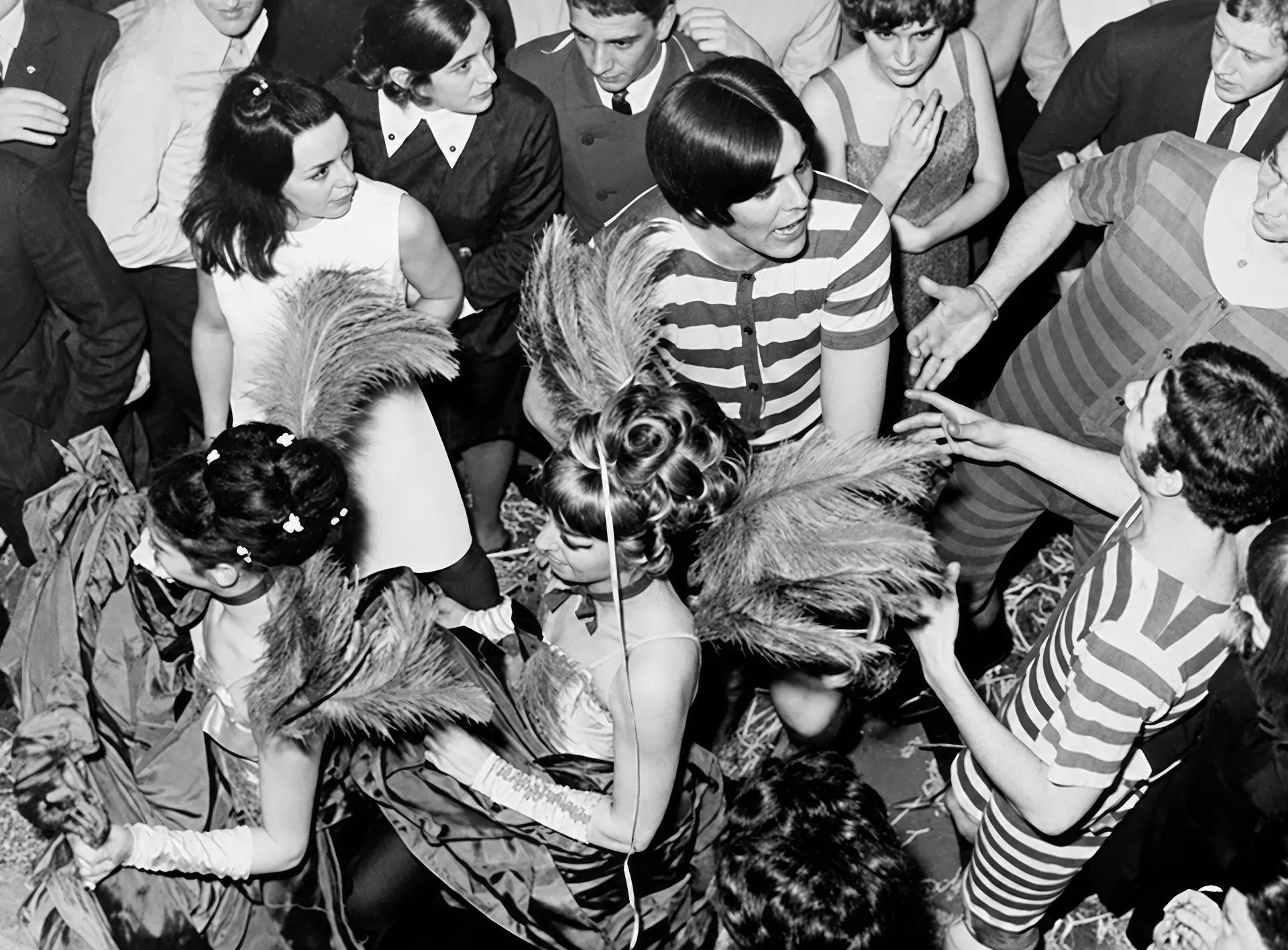 Le public des Gémeaux un soir de fête : plumes, robes à rayures et coiffures crêpées sur la piste couverte de paille. L'idée qu'une soirée se prépare comme un spectacle est déjà là.