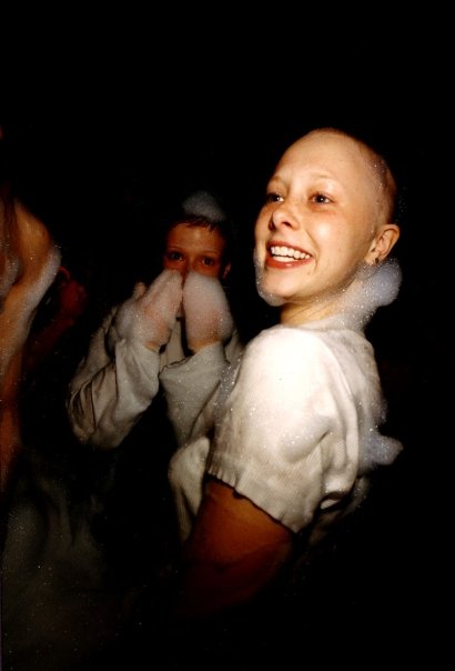 A young woman with a shaved head, beaming, surrounded by foam.