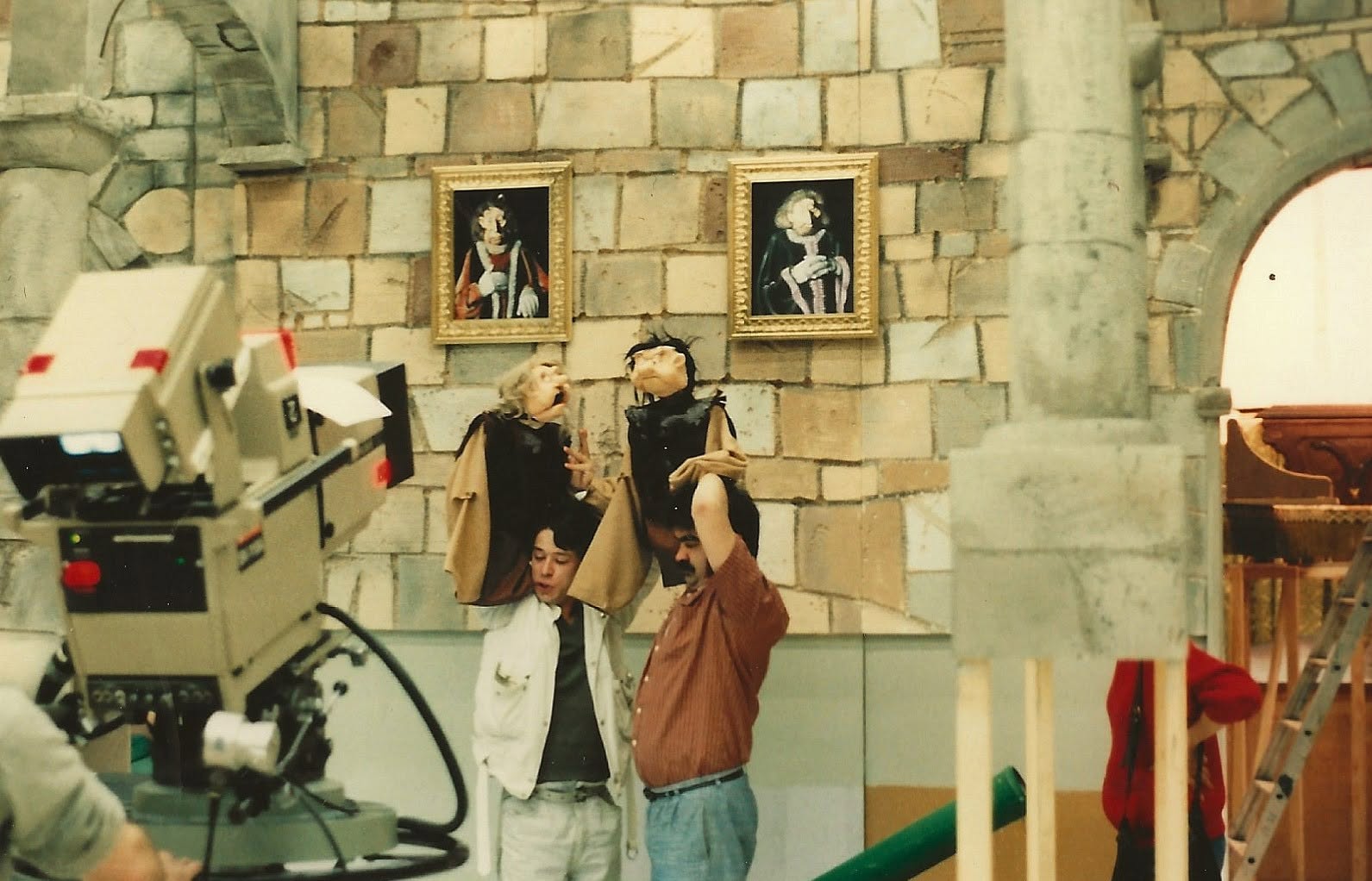 Deux marionnettistes tiennent leurs personnages devant un mur de portraits peints. Le décor va jusqu'aux tableaux accrochés aux murs du château.