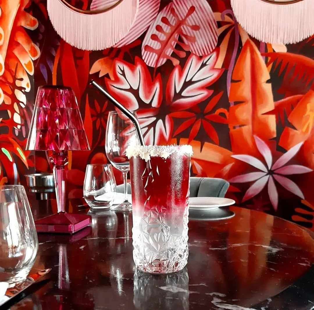 Le détail dit le métier : feuilles découpées au pinceau, dégradés d'orange et de rose, blancs laissés en réserve pour donner la lumière. Au premier plan, un verre taillé et une lampe framboise reprennent exactement les couleurs du mur.