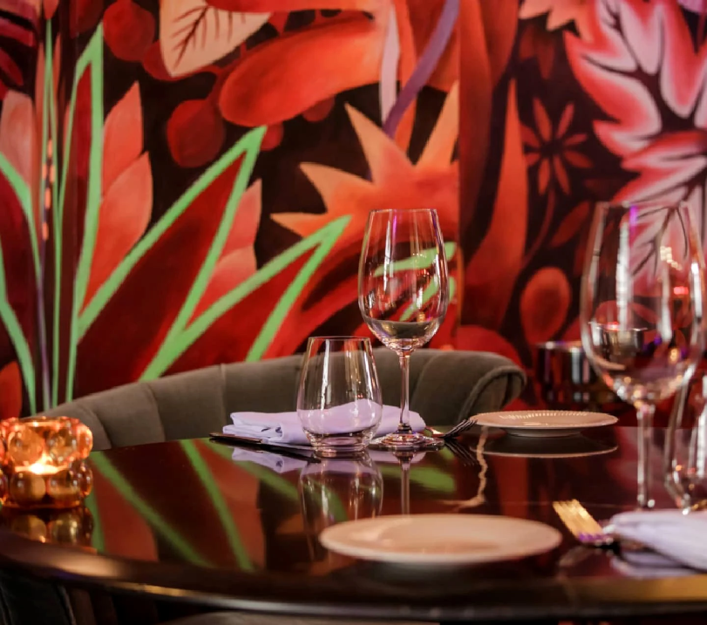 Verres, assiettes et bougie sur le marbre sombre ; derrière, les feuillages rouges et les traits verts de la fresque. Rien ne sépare la table du décor : le mur peint entre dans le service.