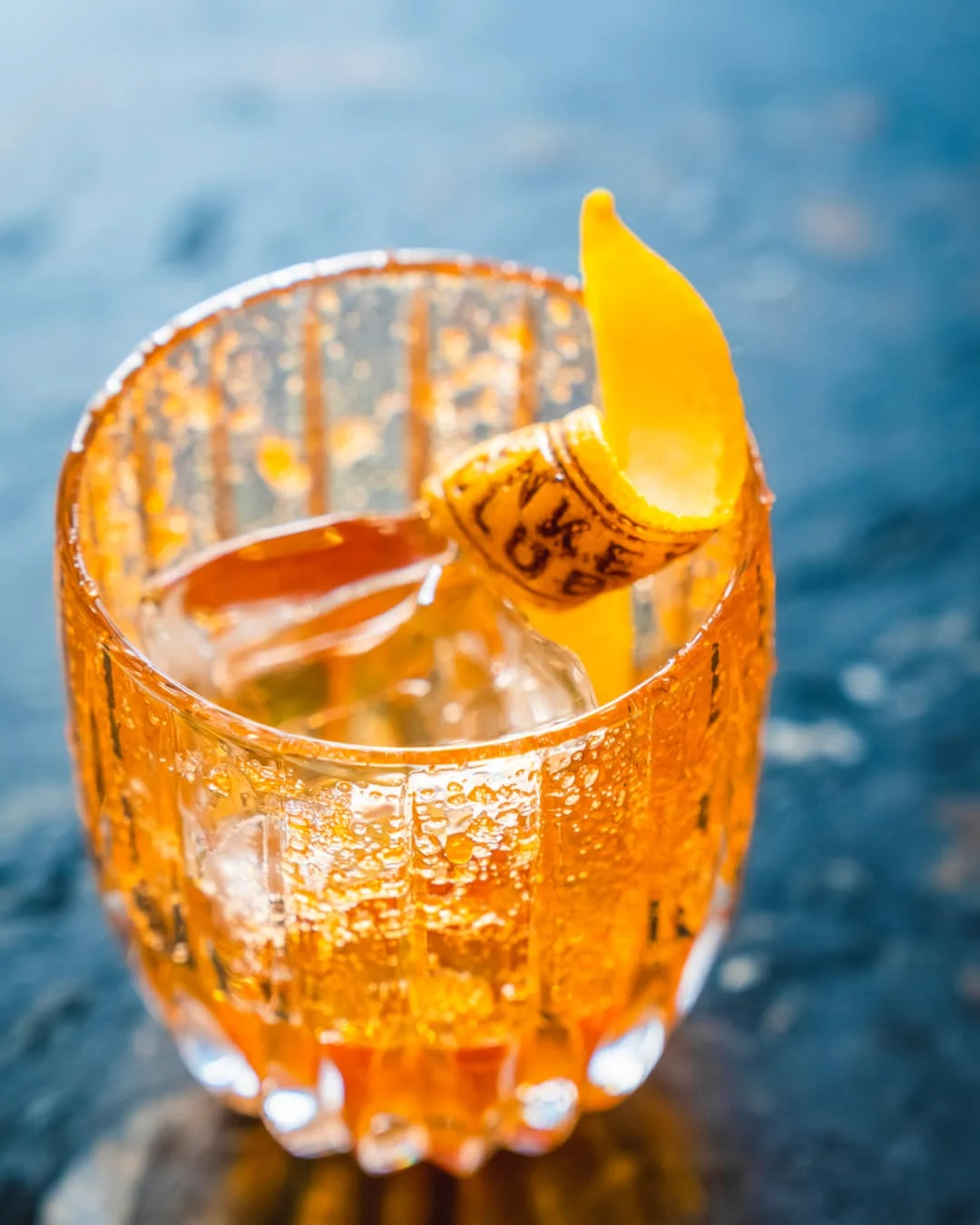 Gros plan sur un verre taillé, glace, bulles et un zeste d'orange posé comme une voile. La photographie de bar tient le même soin du détail que le décor peint.