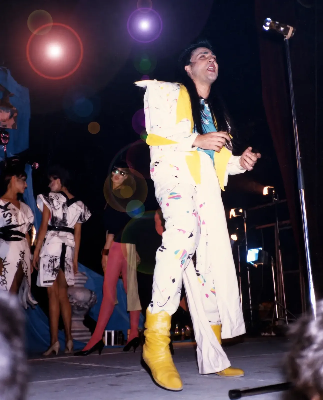 Photographie couleur, format vertical, prise en contre-plongée. Sur la scène du Mirano Continental en 1986, le chanteur, costume blanc peint à la main de motifs jaunes et pastel, bottes jaunes souples et longue mèche noire, se cambre devant le pied de micro. Derrière lui, des mannequins en robes blanches sérigraphiées de dessins noirs attendent leur passage, devant les voilages bleus du décor.