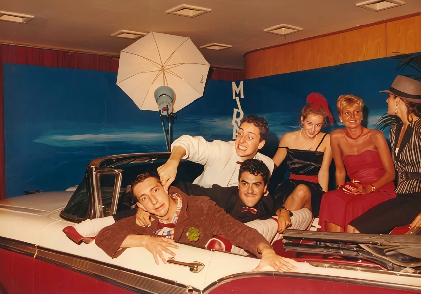 Photographie couleur, format horizontal. Troisième vue de la séance de 1989 : Dan Mac Roll allongé sur la capote de la Cadillac au premier plan, ses complices du Trio Infernal, Santino Del Paradiso Trovato et Riton Liebman, et leurs invitées à l'arrière, devant les lettres géantes du Mirano.