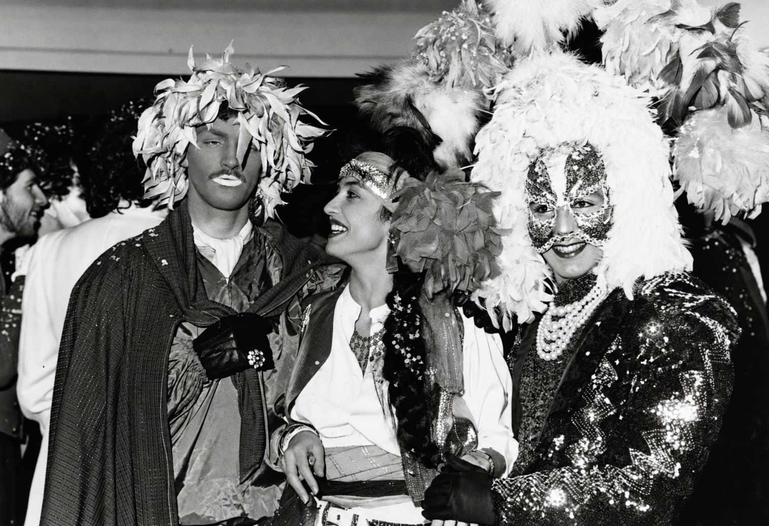Photographie noir et blanc, format horizontal. Un groupe d'habitués du Mirano Continental pose en costumes chamarrés, plumes et masques, dans l'esprit théâtral des soirées à thème du club.