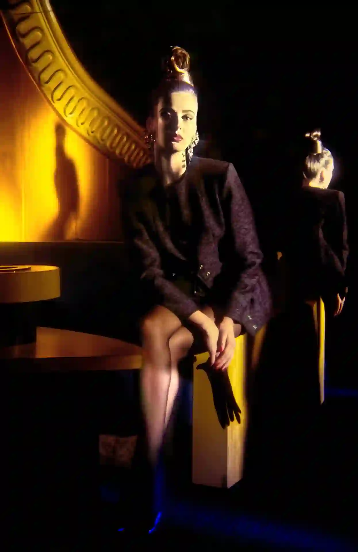 Photographie couleur, format vertical. Dans la lumière dorée du Mirano, une mannequin pose assise sur un socle jaune, veste courte sombre aux épaules carrées, chignon haut doré, boucles d'oreilles pendantes, un gant noir à la main, tandis qu'une seconde silhouette tourne le dos, découpée sur le décor lumineux. En 1987-88, la créatrice Yota Angelaki faisait photographier ses vêtements au Mirano : la salle de nuit servait de studio de pose, entre théâtre et vitrine de mode.