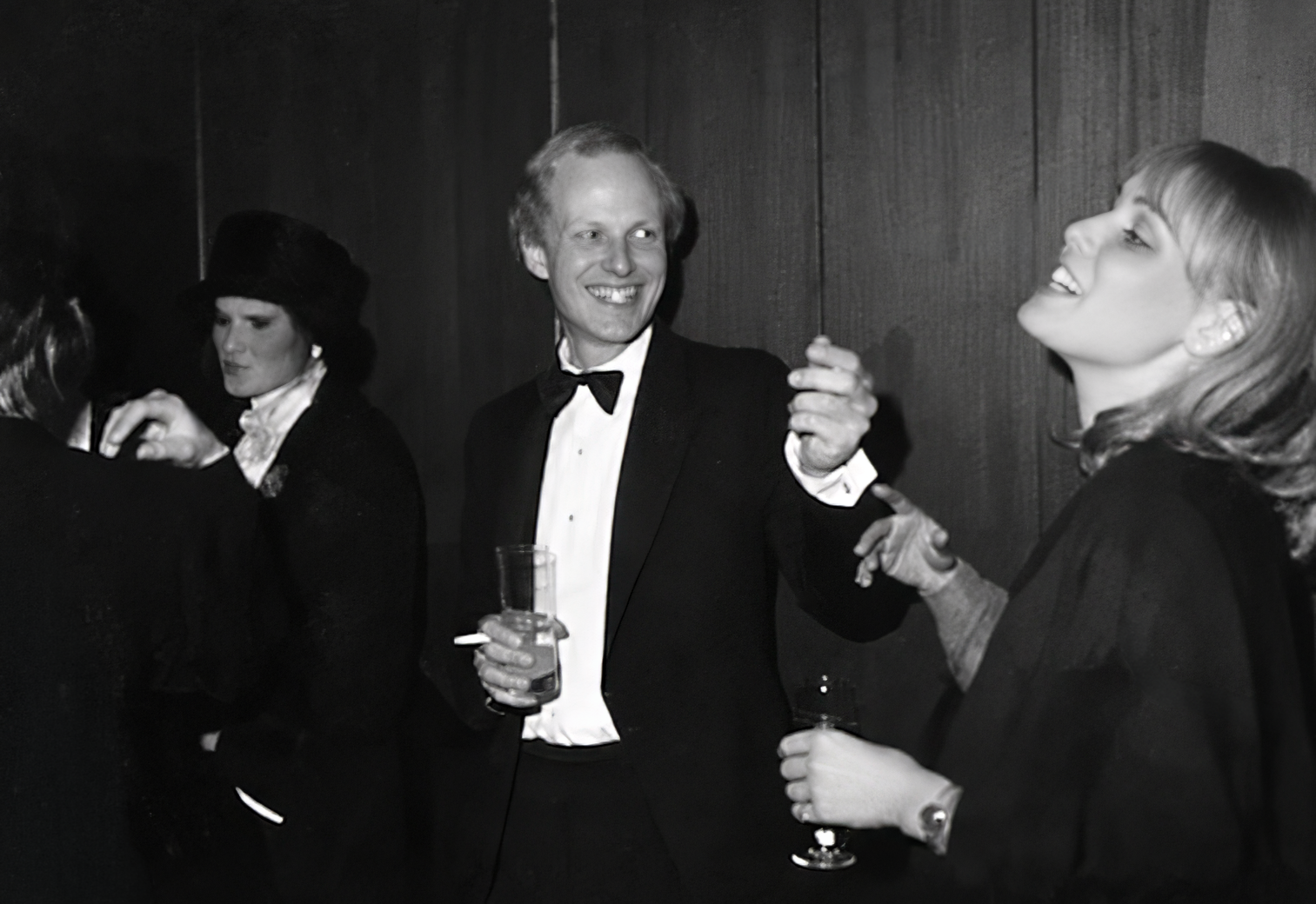Photographie noir et blanc, format horizontal. Au bar du Mirano Continental, un homme en smoking discute avec une femme blonde ; d'autres invités en tenue de soirée complètent l'ambiance feutrée et élégante des nuits mirobolantes.