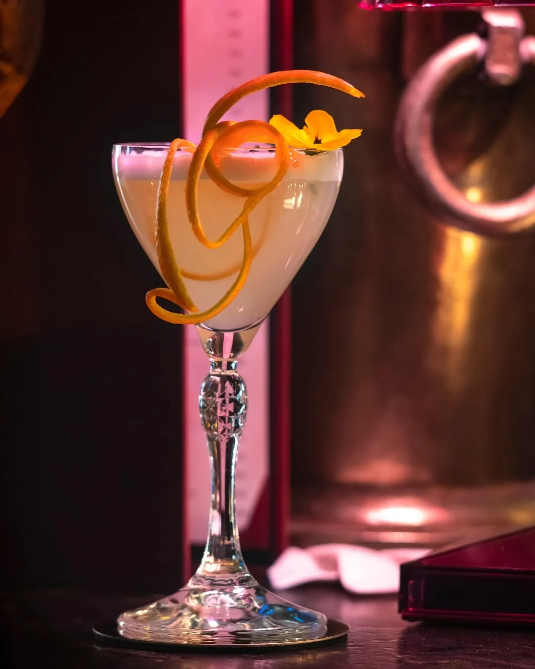 Une coupe ciselée, un zeste d'orange enroulé en spirale et une fleur comestible : la carte des cocktails prolonge le décor, même vocabulaire de couleurs, même goût de la mise en scène.