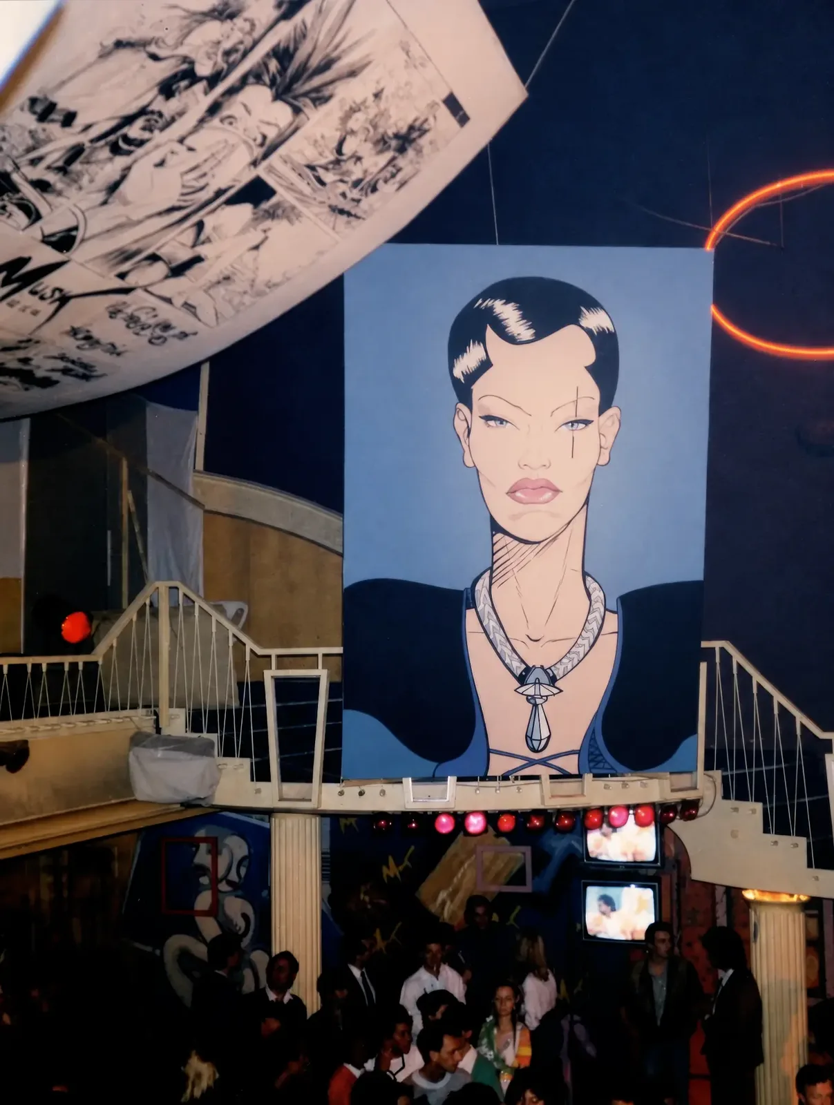 Photographie couleur, format vertical. Au-dessus de la piste du Mirano Continental en 1986, un portrait monumental de Musk, l'héroïne de la bande dessinée, domine la salle : visage dessiné sur fond bleu, frange laquée, collier et décolleté croisé. À gauche, une planche originale géante de l'album est suspendue au plafond ; à droite, un anneau de néon rouge. En contrebas, des écrans de télévision diffusent des images devant le public rassemblé sous les escaliers blancs.