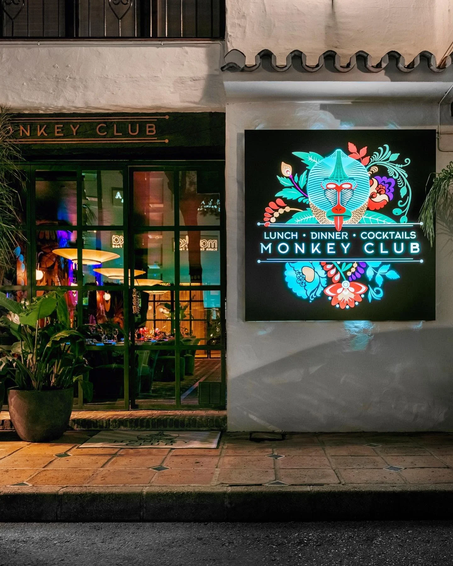 La nuit, le mandrill turquoise s'allume au milieu des fleurs : l'enseigne lumineuse répond, dehors, aux couleurs de la salle. À gauche, les baies vitrées laissent voir les tables dressées sous les grandes coupoles de lumière.