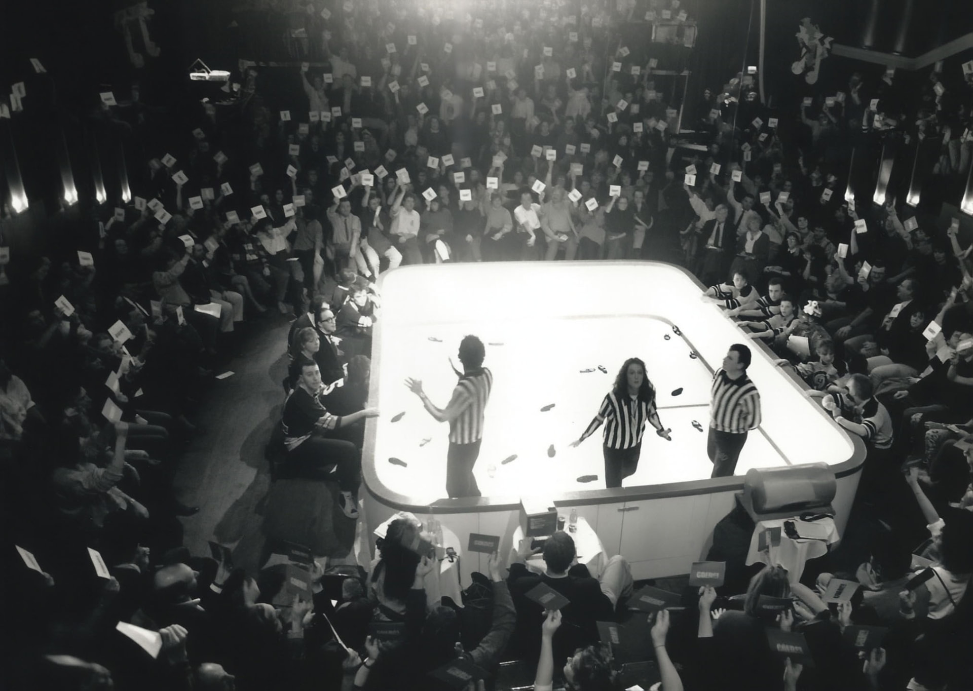 Photographie noir et blanc, format horizontal. Vue en plongée d'un match de la Ligue d'improvisation belge au Mirano Continental : trois comédiens en maillots rayés d'arbitre sur la patinoire centrale éclairée, entourés sur quatre côtés par environ 500 spectateurs brandissant leurs cartons de vote, dans une ambiance de véritable chaudron.