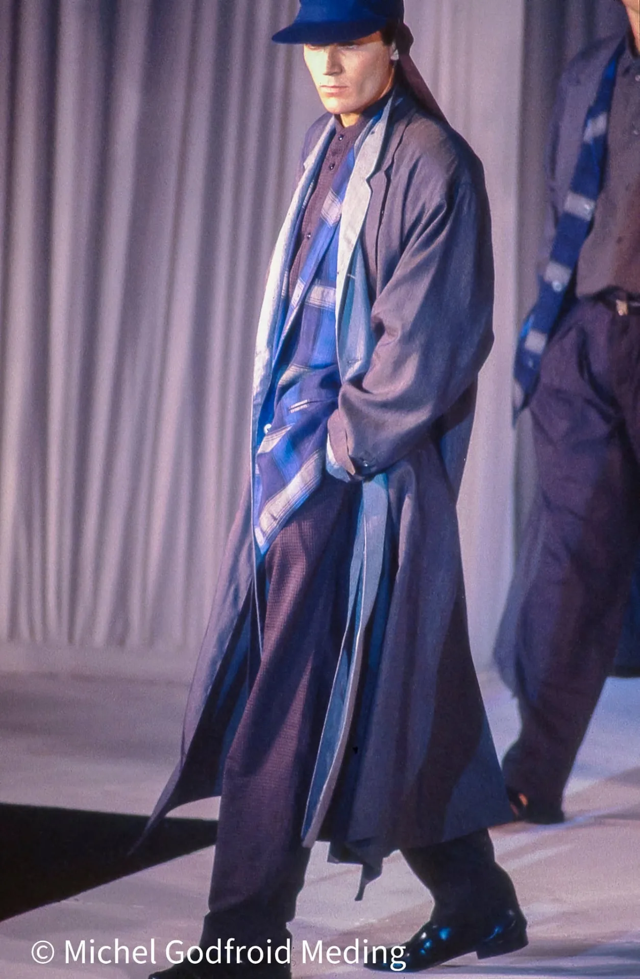 Photographie couleur, format vertical. Un mannequin traverse le podium, long manteau gris fluide porté sur des chemises à carreaux bleutés, chapeau assorti, mains dans les poches. La superposition des couches et le jeu des bleus donnent à la silhouette une élégance de bande dessinée.