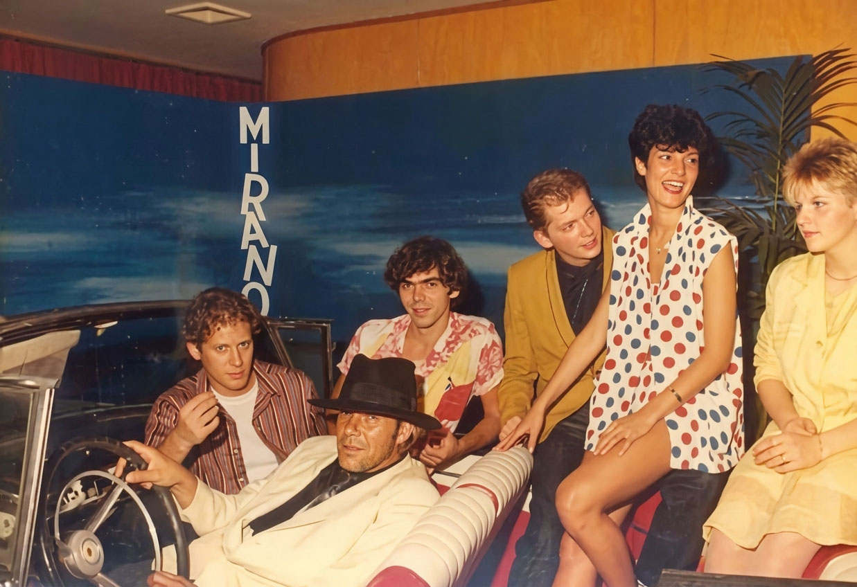 Photographie couleur, format horizontal. Même séance de 1989 : un homme en costume blanc et chapeau noir au volant de la Cadillac blanche, entouré d'invités et du logo vertical Mirano dans le hall d'entrée du club.