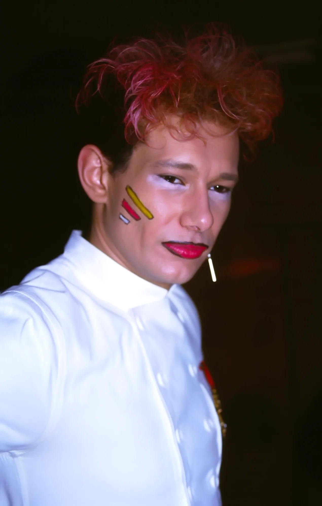 Photographie couleur, format vertical. Portrait de Nanard lors d'une soirée fluo du Mirano Continental : chevelure rouge ébouriffée, trois traits jaune, rouge et bleu peints sur la joue, bouche carmin et chemise blanche immaculée. Années 1980.