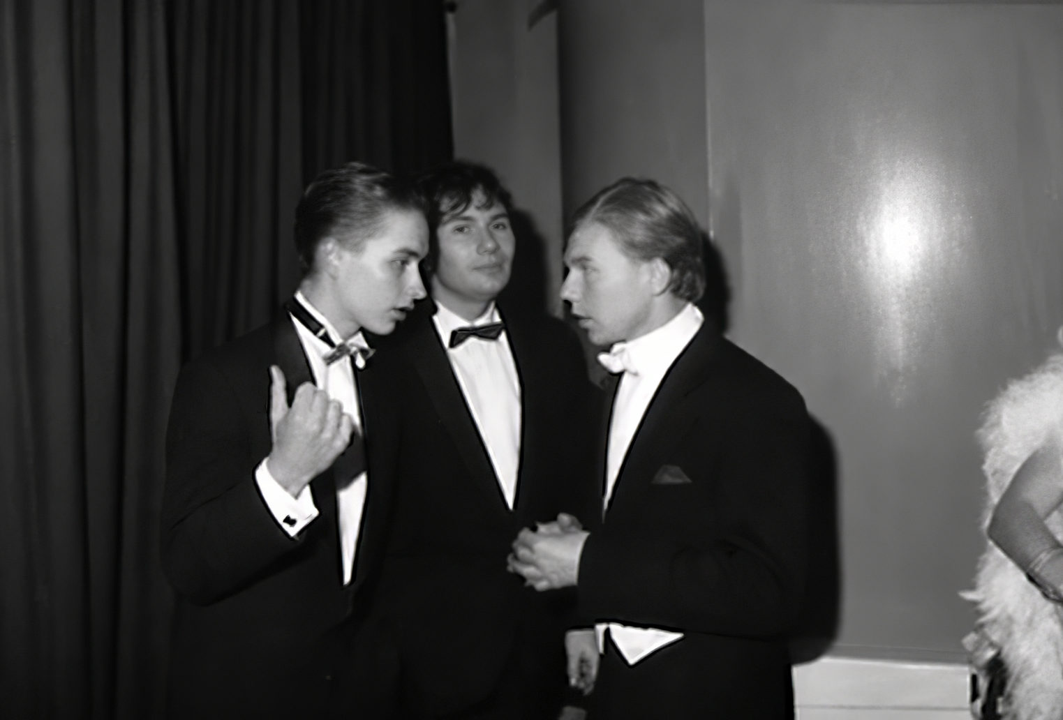 Photographie noir et blanc, format horizontal. Trois hommes en smoking sont rassemblés au bar du Mirano Continental, l'un d'eux faisant un geste de la main, dans la conversation animée de la nuit.
