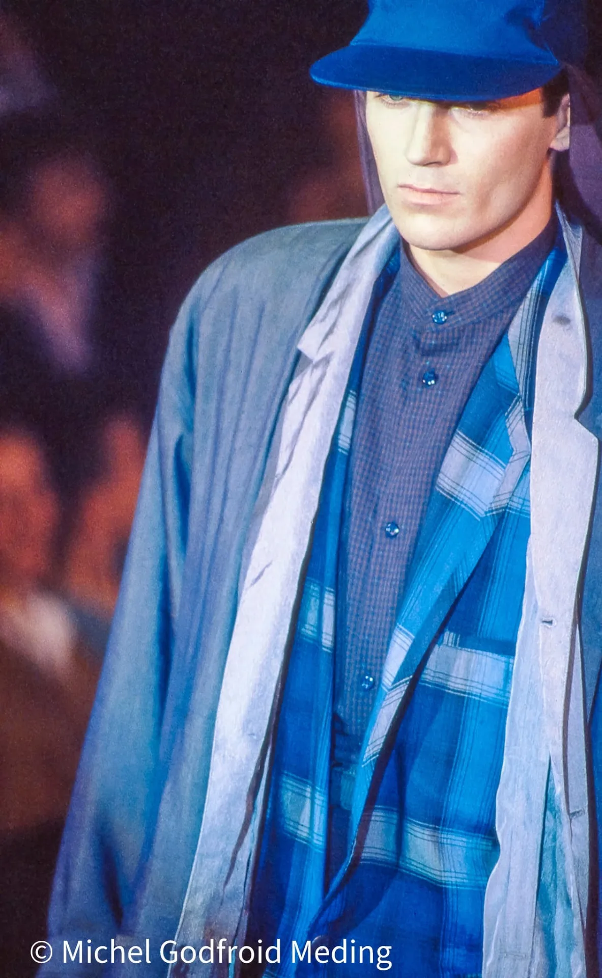 Photographie couleur, format vertical. Un mannequin avance, chapeau mou bleu roi rabattu sur le regard, chemises à carreaux superposées dans tous les tons de bleu sous une veste claire. Prise au plus près, l'image capte l'allure du défilé : le vestiaire devient silhouette, dessiné par la lumière bleutée de la scène.