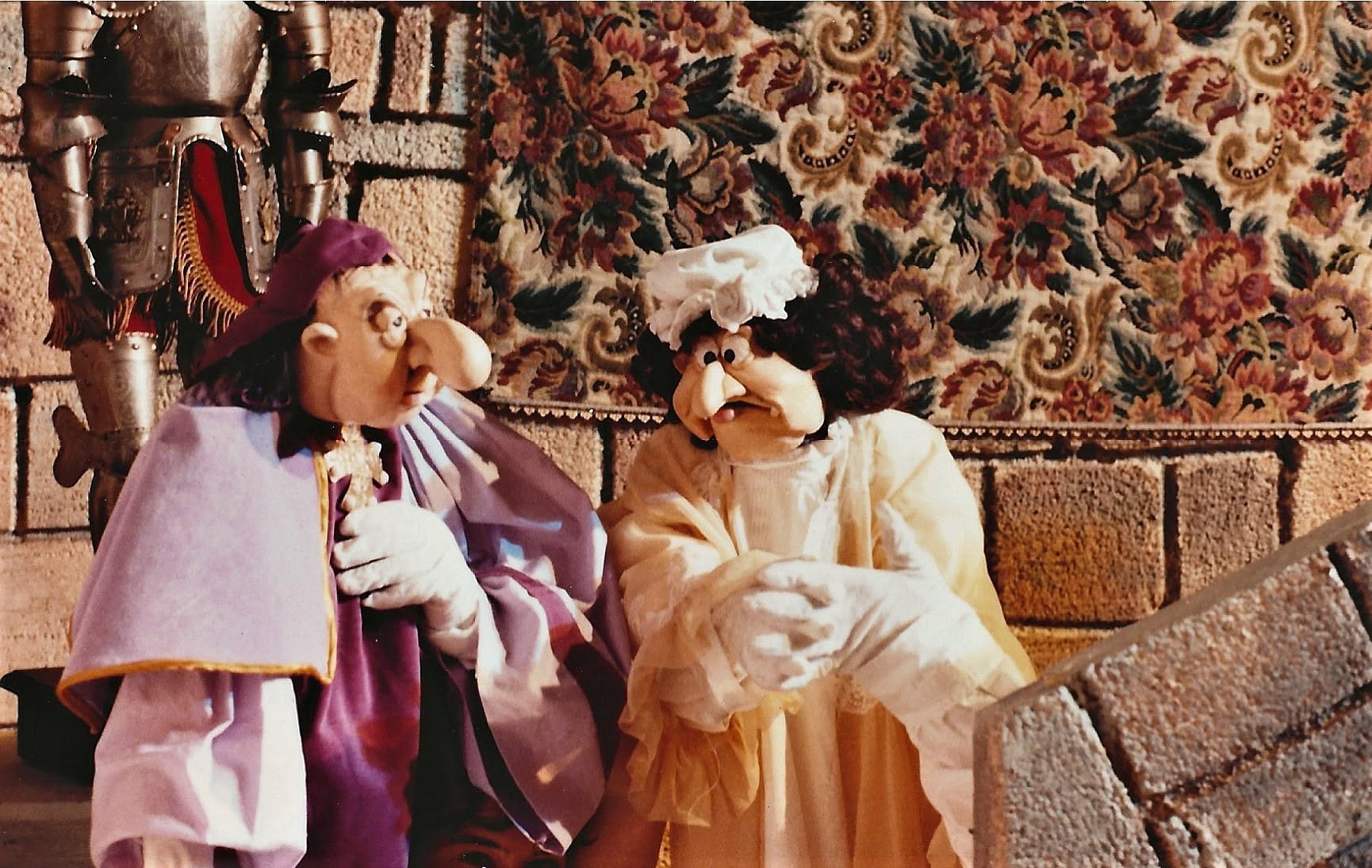 Deux marionnettes en costume devant une tapisserie et une armure : le soin apporté aux visages sculptés et aux étoffes.