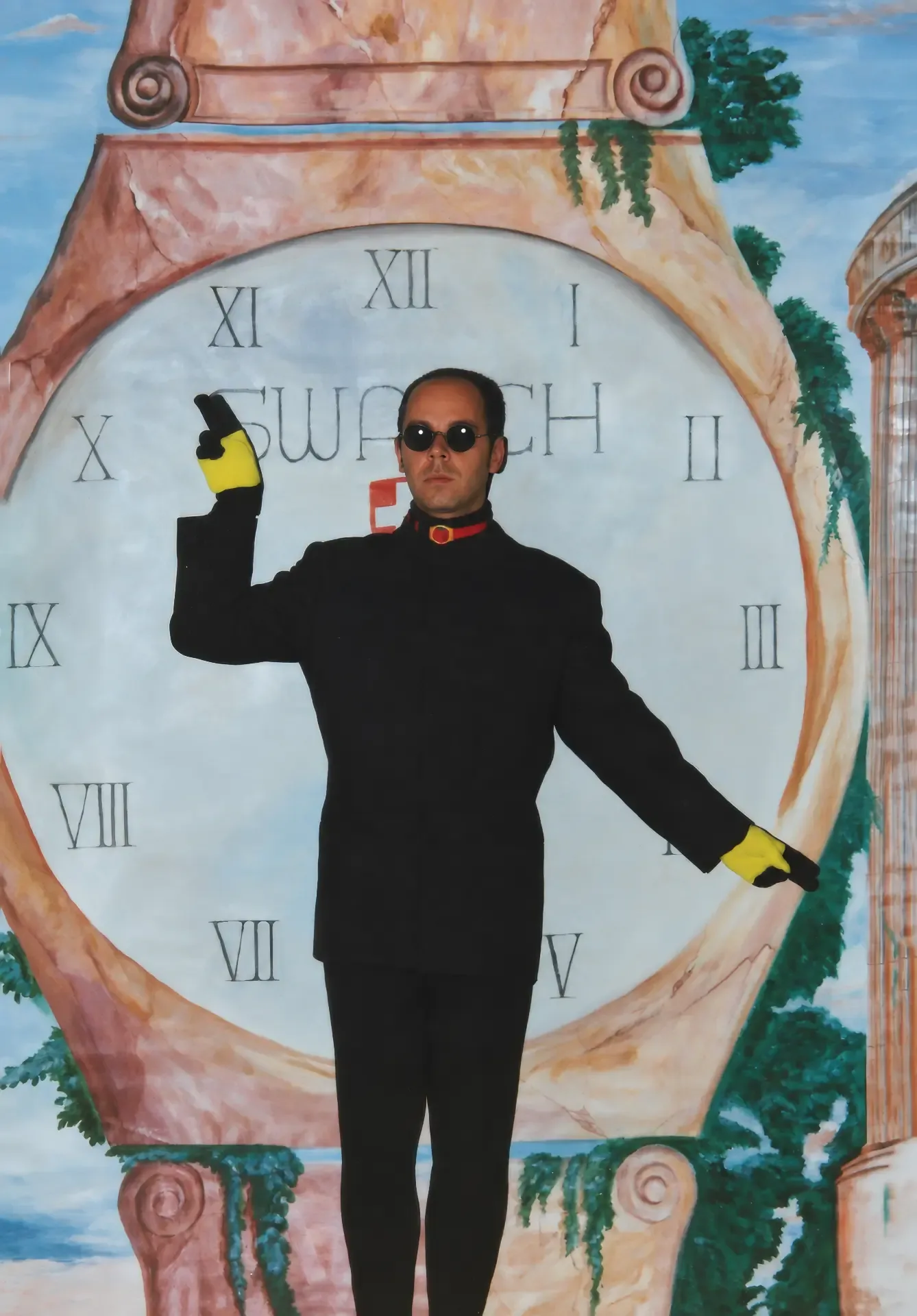 Photographie couleur, format vertical. Devant une toile peinte à la main, un cadran de montre aux chiffres romains encadré de colonnes de marbre en trompe-l'œil, de lierre et de ciel bleu, un performeur en costume noir, lunettes rondes noires et gants jaunes mime les aiguilles, un bras levé, l'autre tendu à l'horizontale. Le décor peint et le corps du performeur ne font qu'un : la marque devient tableau vivant.