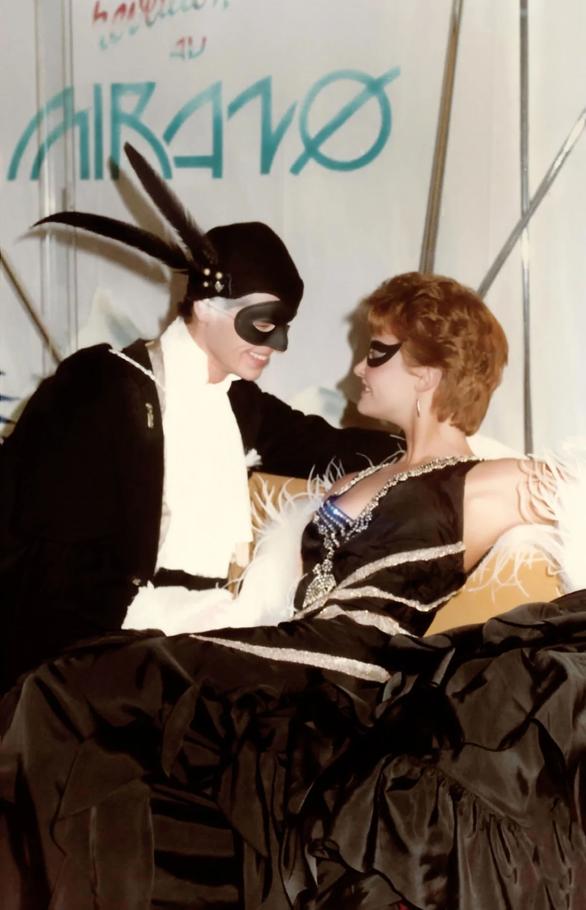 Photographie couleur, format vertical. Deux invités en costumes de bal masqué se font face sous le logo turquoise du Mirano Continental : l'homme porte un loup noir, un chapeau à plumes et une cape blanche ; la femme, robe noire à volants et plumes blanches, lui répond dans un masque assorti. L'image saisit le rapport de séduction propre aux soirées costumées du club au début des années 1980.
