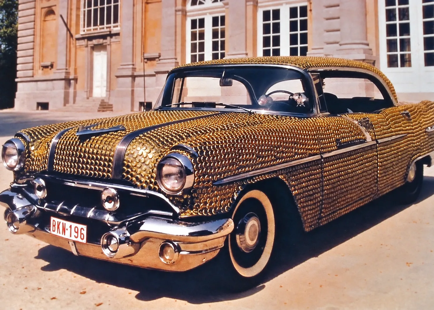 Photographie couleur, format horizontal, prise devant une façade classique en pierre. La carrosserie, capot, ailes, portières et toit, disparaît sous des milliers de capsules dorées posées une à une, en rangs serrés qui épousent les courbes ; seuls les chromes, les pare-chocs et les vitres restent nus. Plaque belge BKN 196. De près, l'effet tient de l'écaille et de la mosaïque : un travail d'artisan appliqué à une opération publicitaire.
