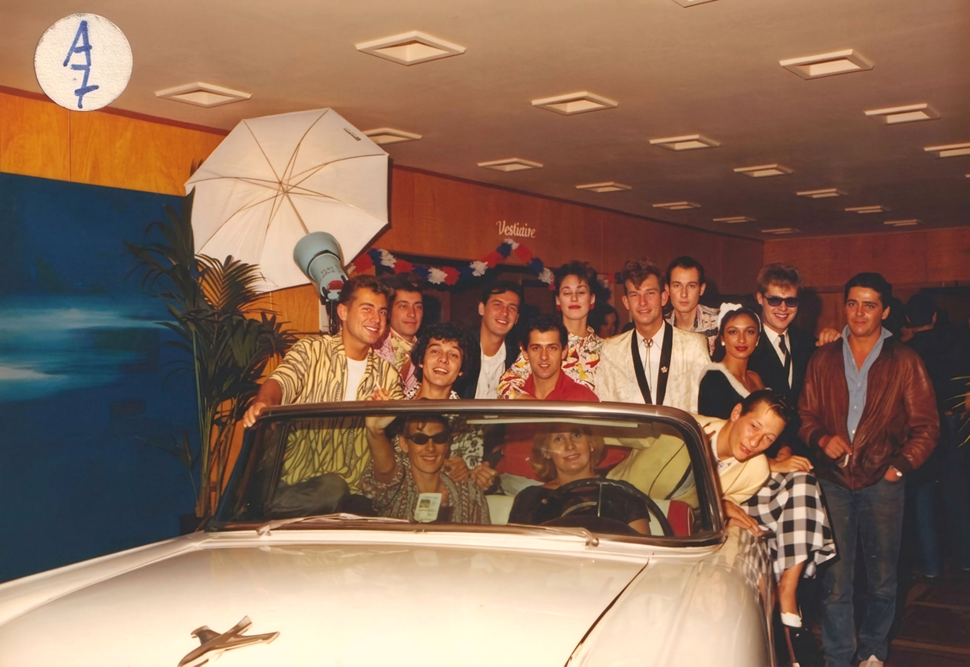 Photographie couleur, format horizontal. Autre vue de la séance de 1989 dans le hall d'entrée du Mirano Continental : la Cadillac blanche, installée comme point de rendez-vous, est entourée d'invités sous le parapluie réflecteur du studio photo et devant le décor bleu nuit au logo vertical du club.