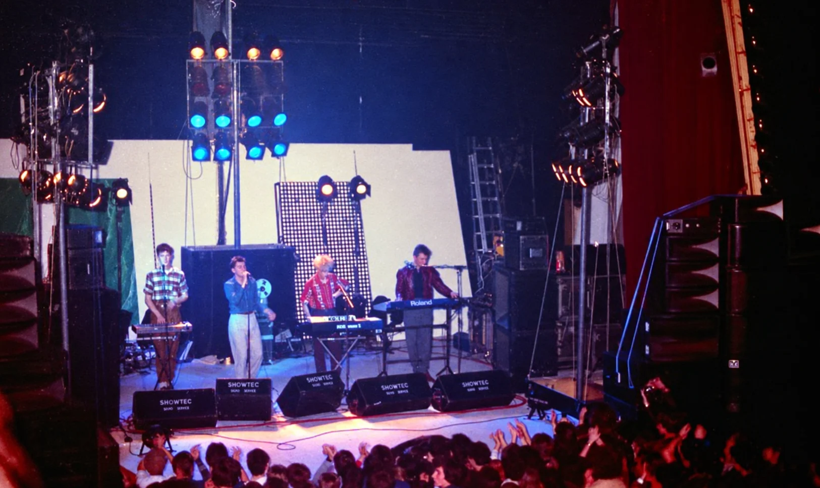 Vue d'ensemble de la scène du Mirano pendant le concert de Depeche Mode, dimanche 12 décembre 1982 : le groupe aligné derrière ses claviers Roland et Korg, panneau lumineux et projecteurs de couleurs à l'arrière, retours Showtec au bord du plateau et public serré au premier rang.