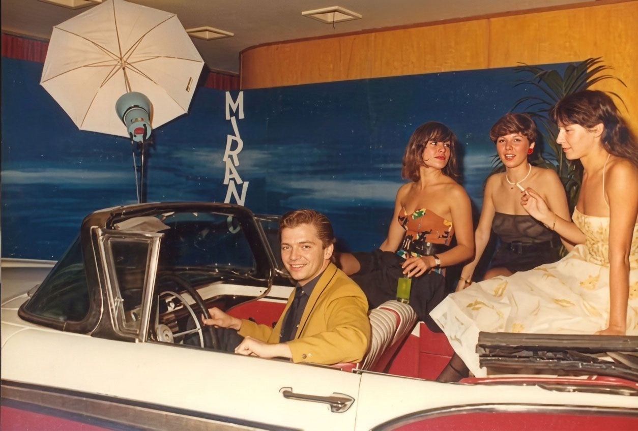 Photographie couleur, format horizontal. Un homme en veste moutarde au volant de la Cadillac blanche, trois femmes élégantes posant à bord, sous le parapluie réflecteur du studio photo et le décor bleu nuit du Mirano Continental. 1989.