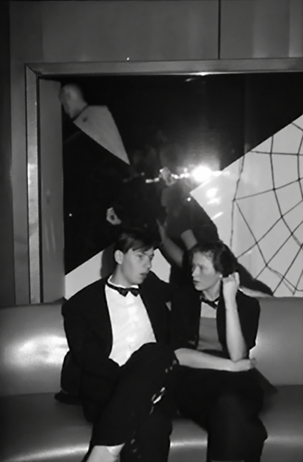 Photographie noir et blanc, format vertical. Un homme en smoking est assis sur le canapé du Mirano Continental, devant un grand miroir aux motifs géométriques qui renvoie les lumières de la salle.