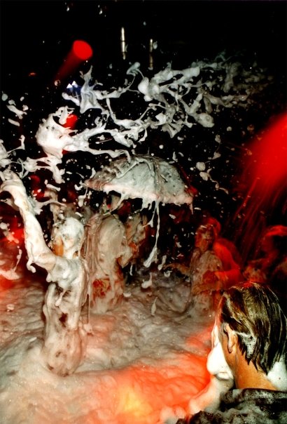 Foam bursting above the dance floor, an open umbrella in the crowd, under red spotlights.