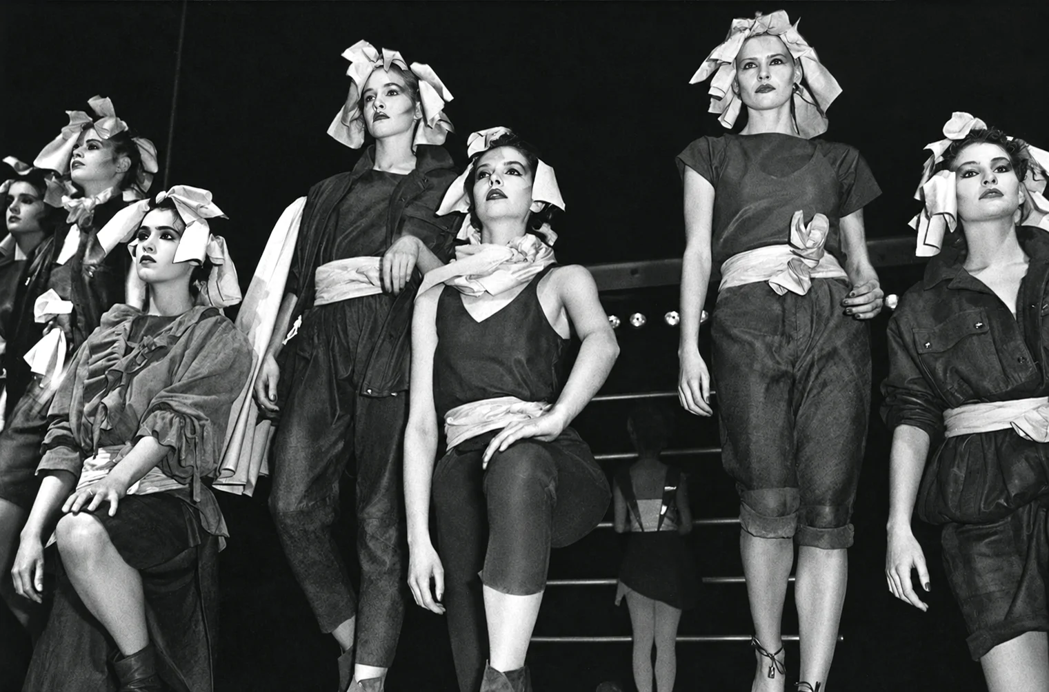 Photographie noir et blanc, format horizontal. Sur la scène du Mirano, sept mannequins composent un tableau final : tissus noués en turban, ceintures drapées à la taille, vestes oversize et pantalons courts, entre influence japonaise et allure de bohème. Les corps se tiennent comme sur une scène de théâtre, visages levés vers la lumière.