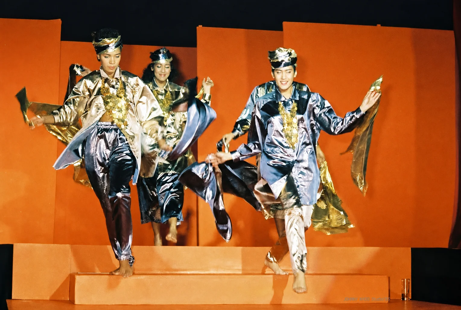 Photographie couleur, format horizontal. Devant un mur orange vif, trois danseurs-mannequins descendent un escalier en lamés argent et or, casquettes brillantes et colliers de sequins, pieds nus, étoffes déployées en mouvement. Le défilé devient danse : la mode au Mirano se jouait avec l'énergie d'un spectacle, dans les couleurs franches qu'Aldo Gigli aimait donner à ses décors.