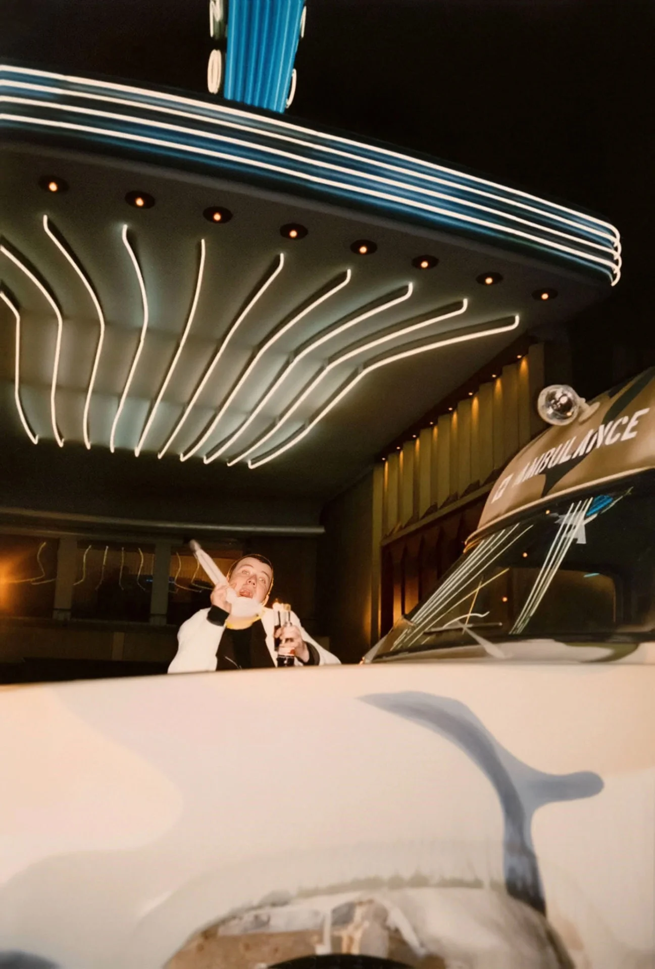 Photographie couleur, format vertical. Jan Bossuyt, figure de l'équipe du Mirano Continental, pose devant l'entrée du club illuminée de néons bleus, un verre à la main, aux côtés d'une ambulance customisée pour une soirée. 1994.